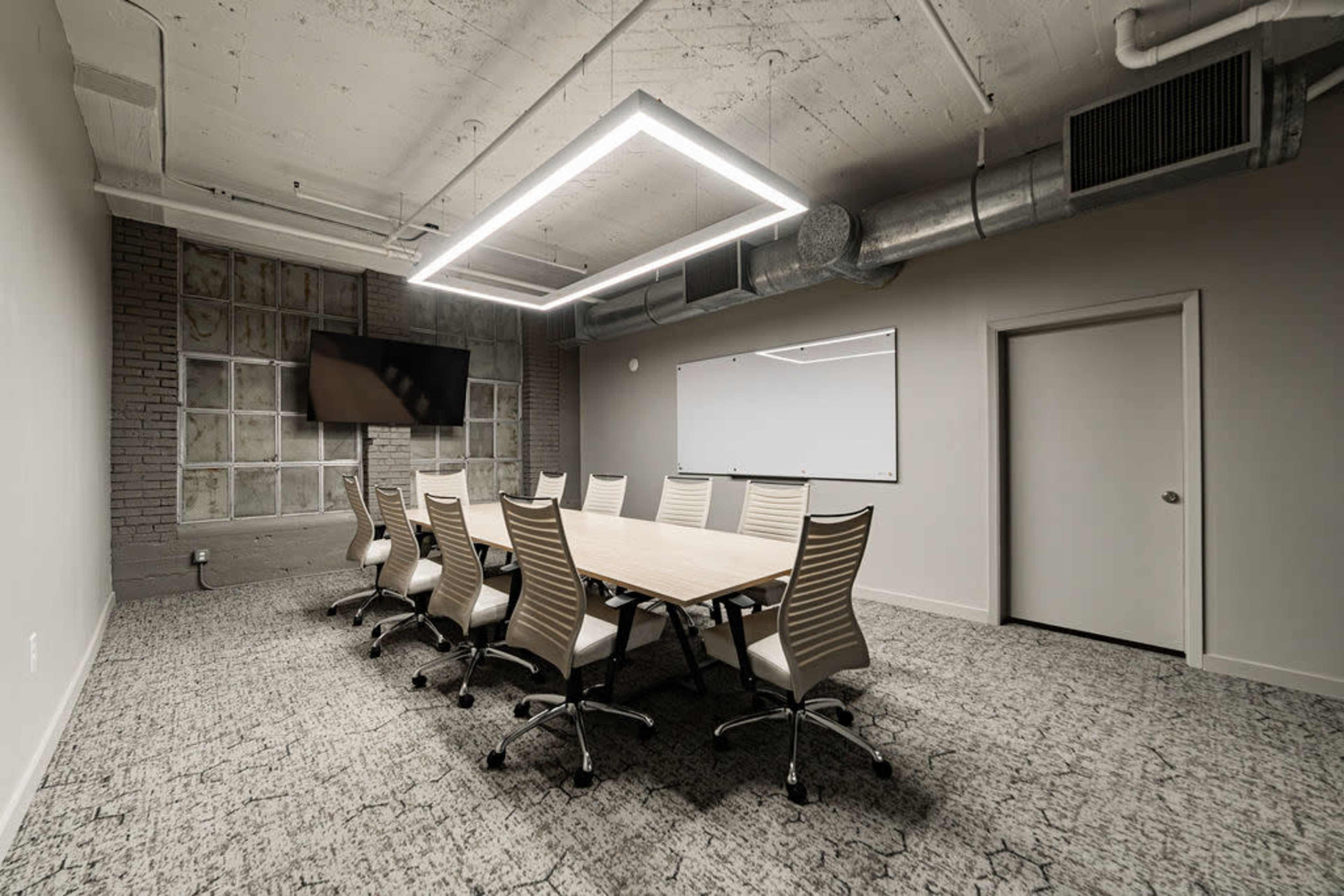 A modern conference room features a long wooden table surrounded by white ergonomic chairs, with a large wall-mounted screen and a whiteboard.