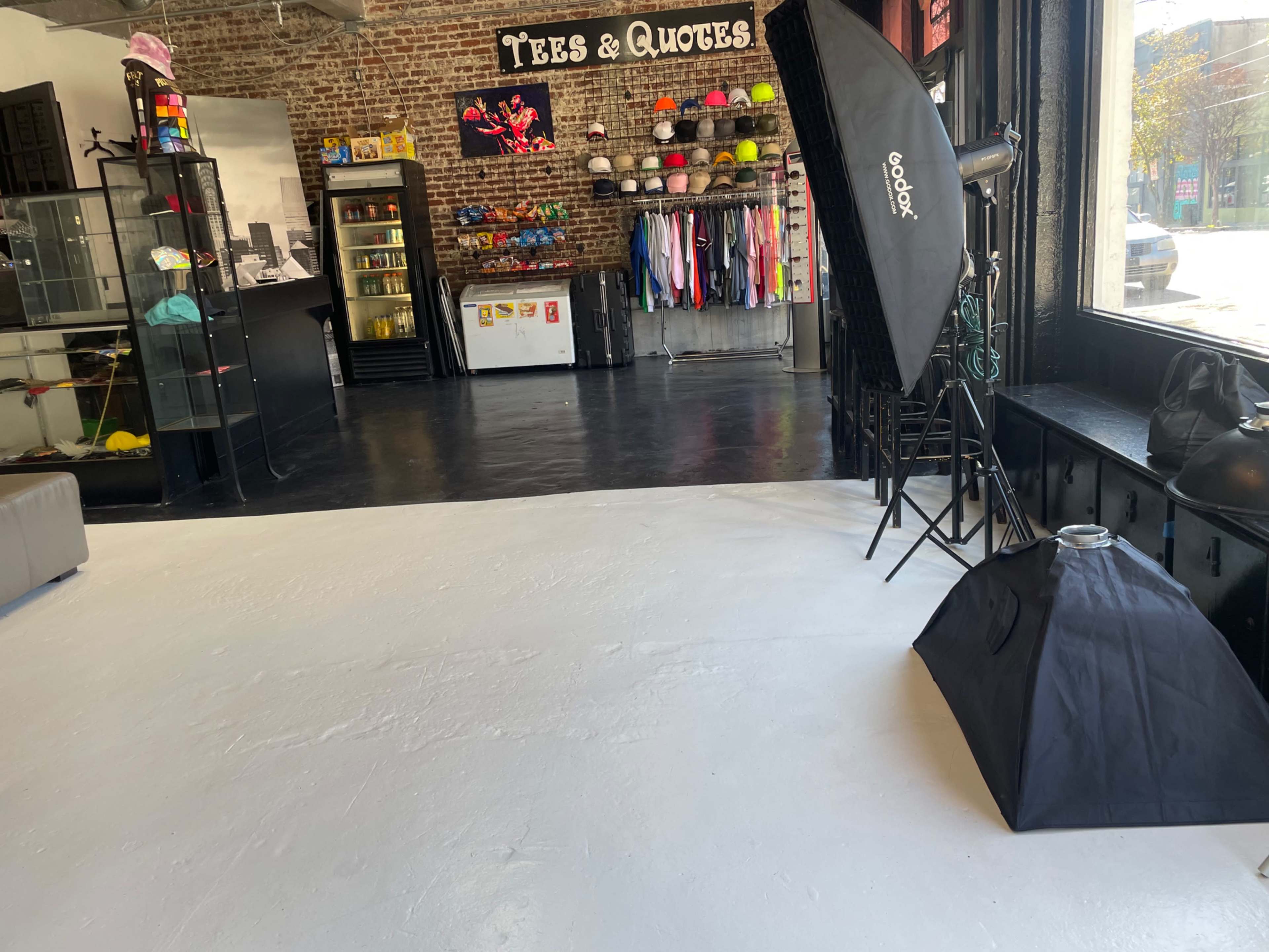 A retail space with exposed brick walls features clothing racks, a display case, and photography equipment set up on a white floor.