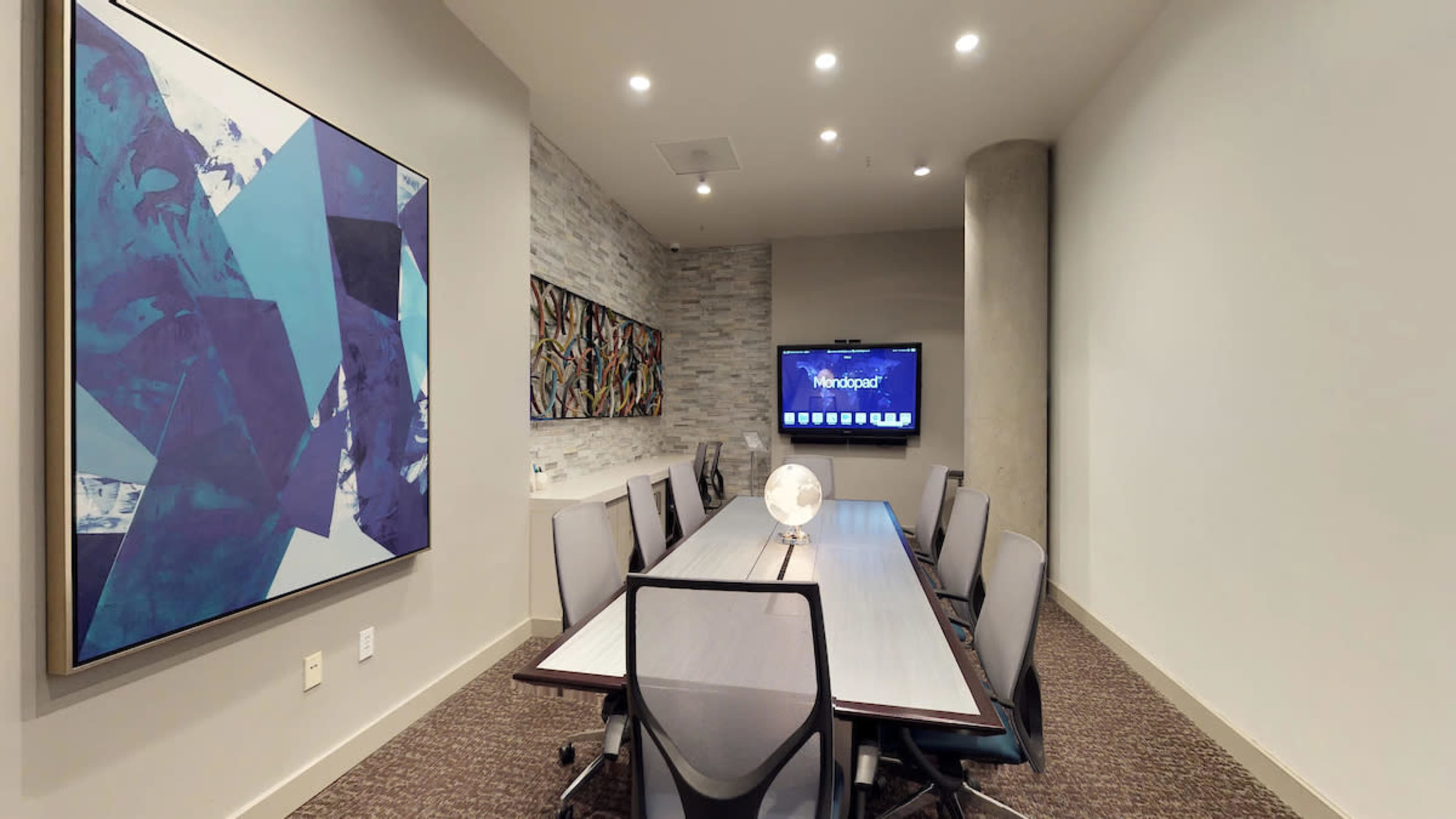 A modern conference room with a long table, several chairs, a large wall-mounted screen, and abstract artwork on the walls.