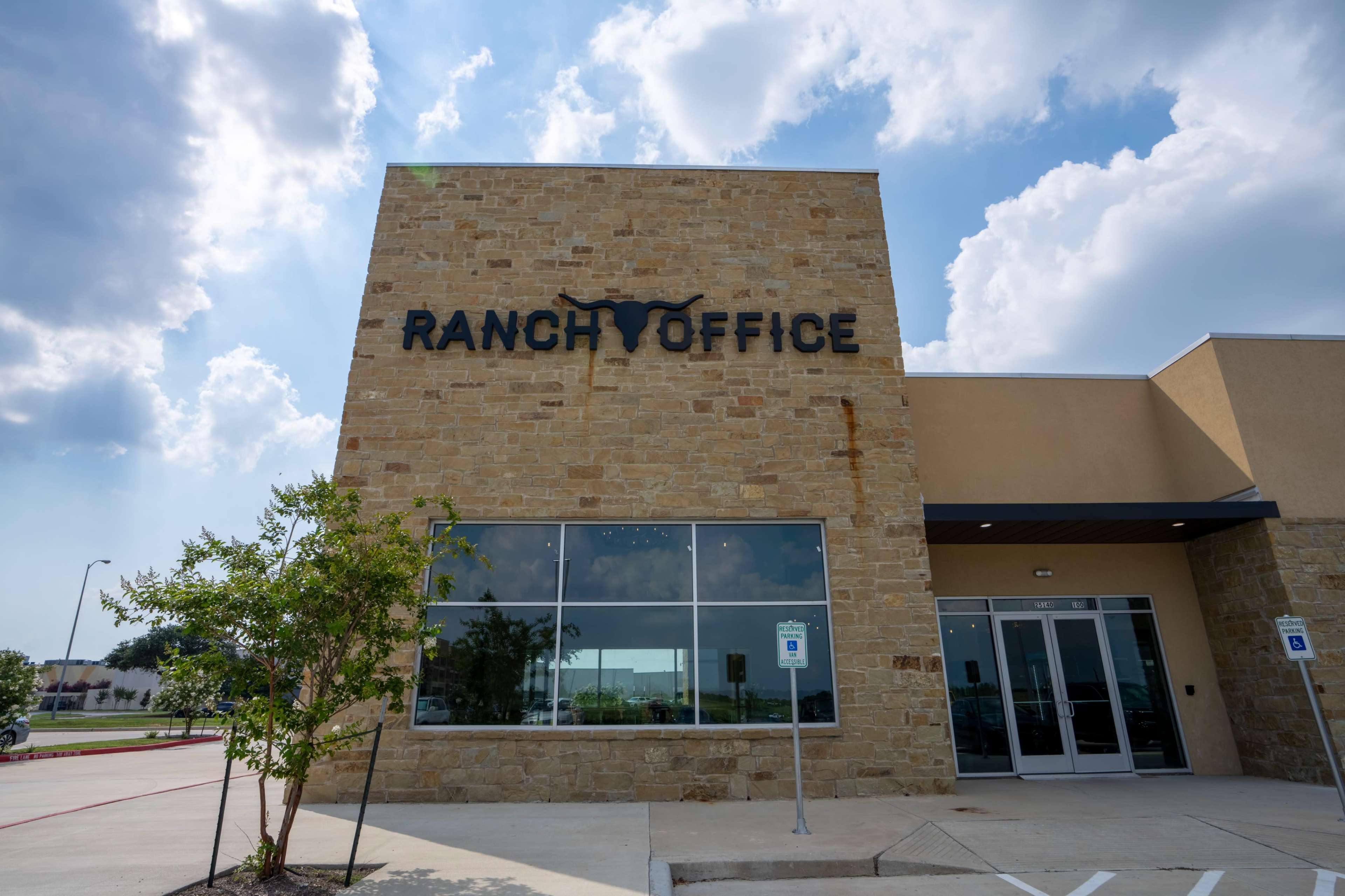 A stone building with large lettering that reads "RANCH OFFICE" and features multiple windows under a partly cloudy sky.