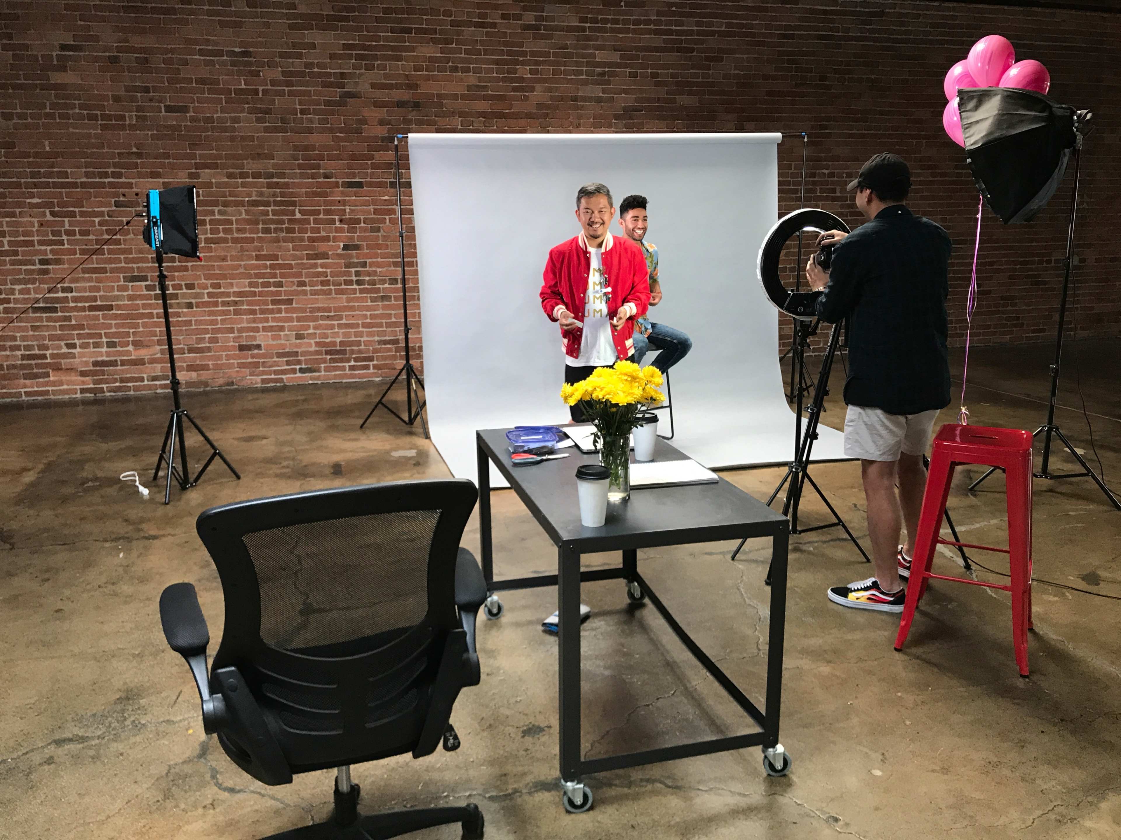 A photo shoot is taking place in a studio with a backdrop, featuring a man in a red jacket and another person in the background, while a third person adjusts lighting equipment.