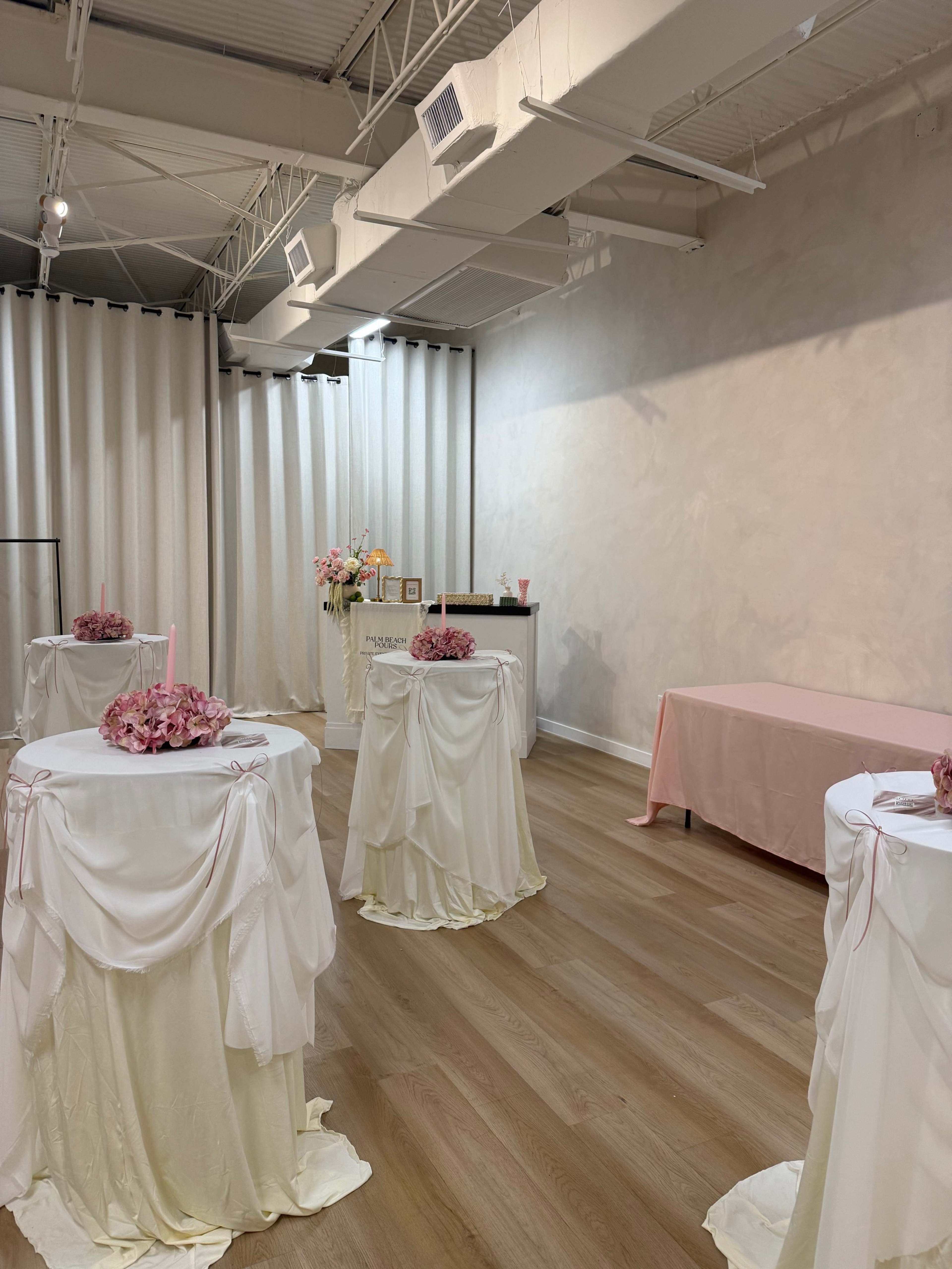 The image shows a light-colored room with draped tables adorned in white and pink fabric, arranged for an event.