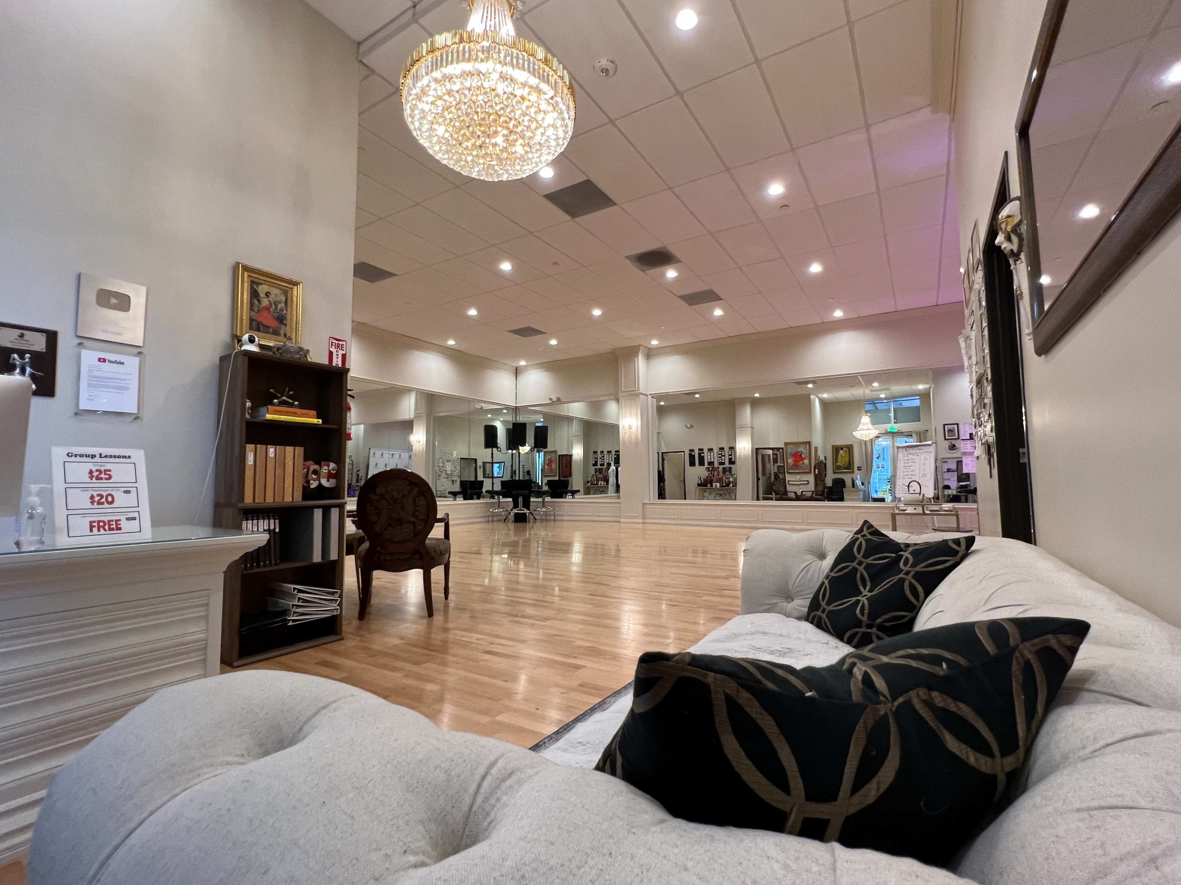 A spacious dance studio with wooden flooring, mirrors on the walls, and a chandelier overhead, featuring a comfortable couch and a decorative chair in the foreground.