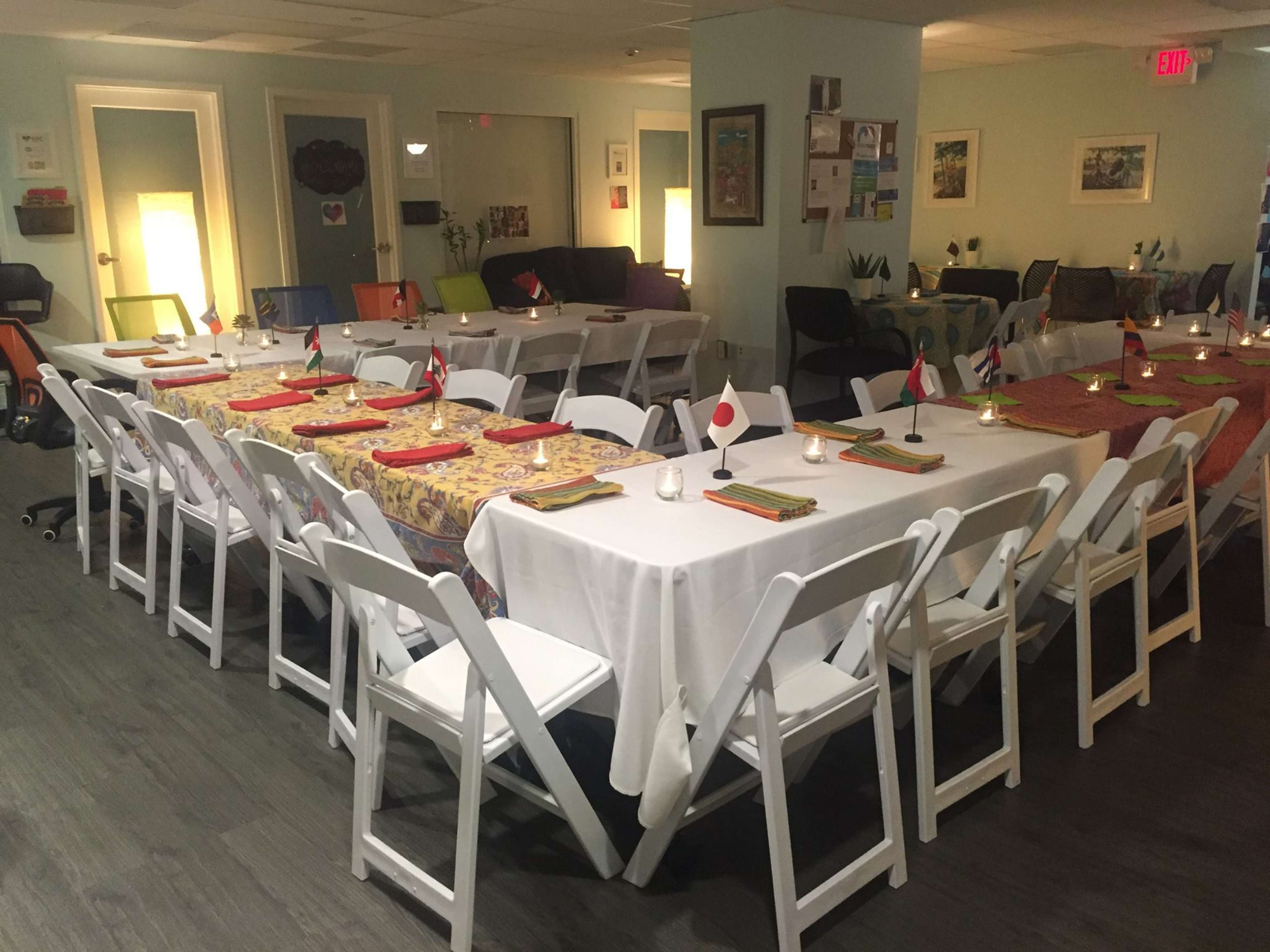 A dining area is set up with multiple tables arranged in a U-shape, adorned with tableware and flags from various countries, ready for a gathering.