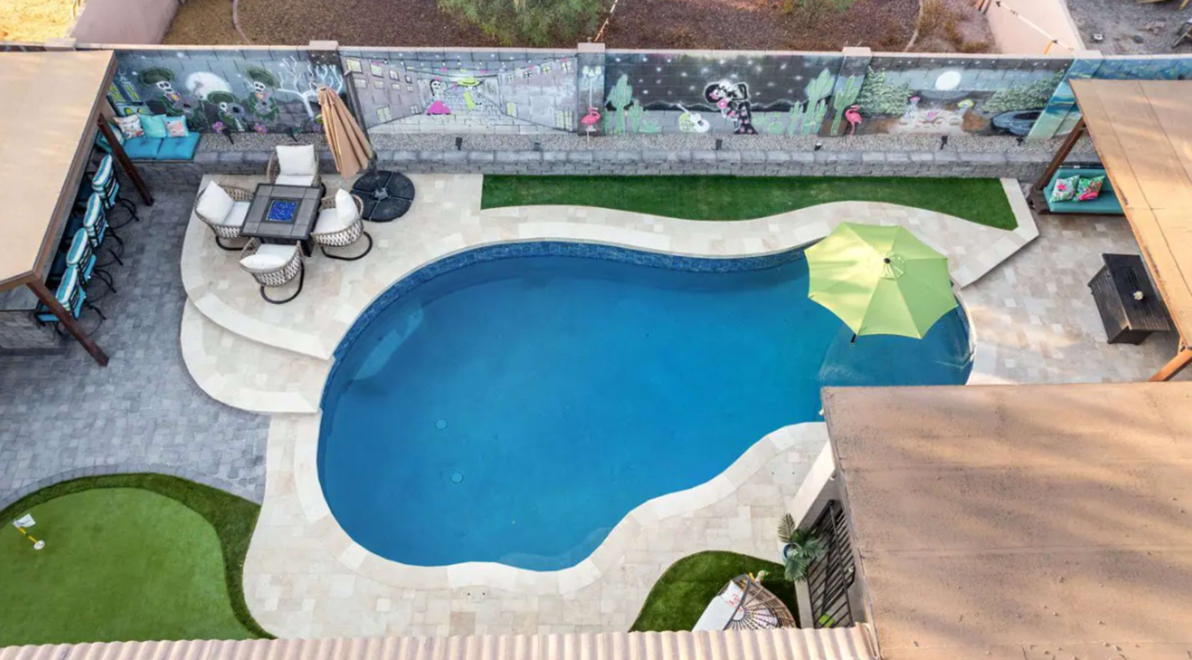 An aerial view of a swimming pool surrounded by a patio, lounge chairs, and an umbrella, with a mural-covered wall in the background.