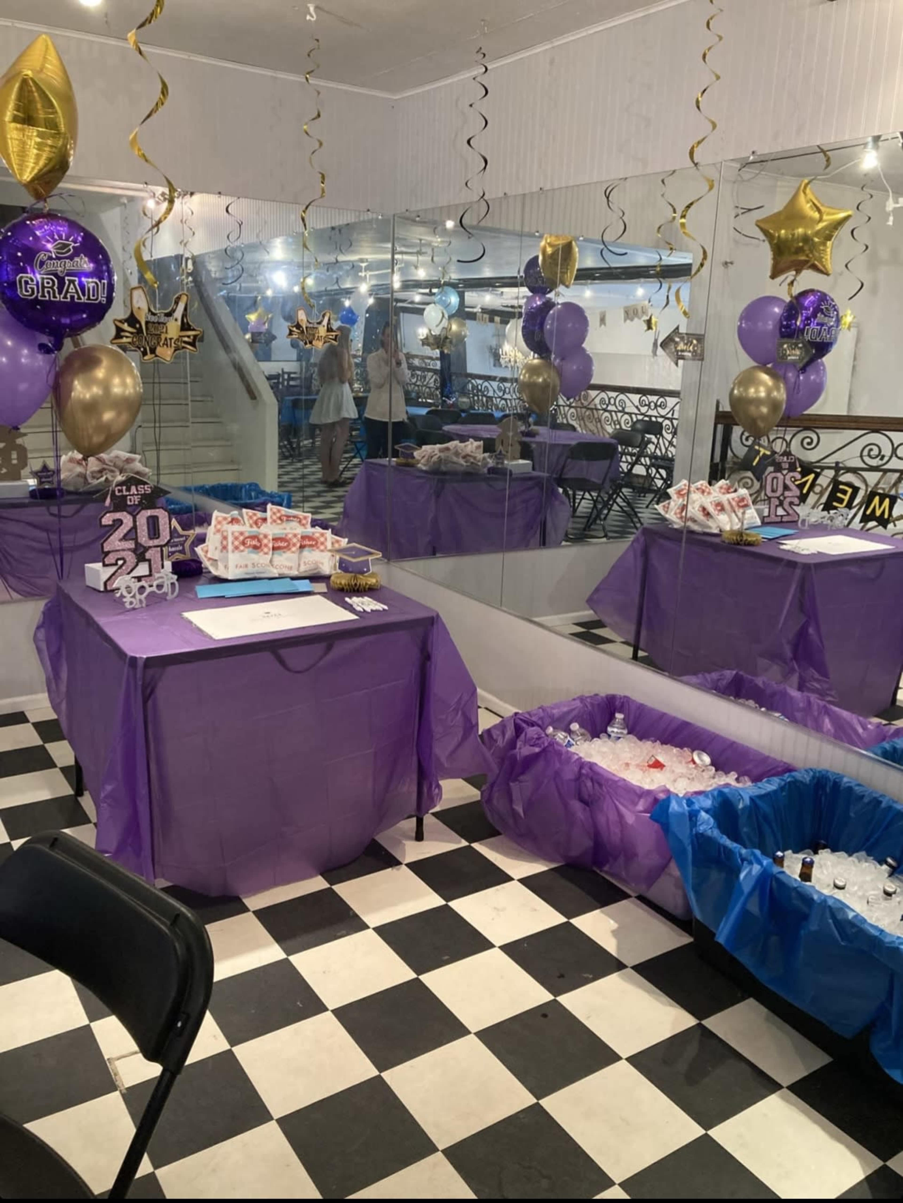 The scene depicts a decorated celebration area featuring purple and gold balloons, a table with gift boxes, and a mirrored wall, set with a black and white checkered floor.