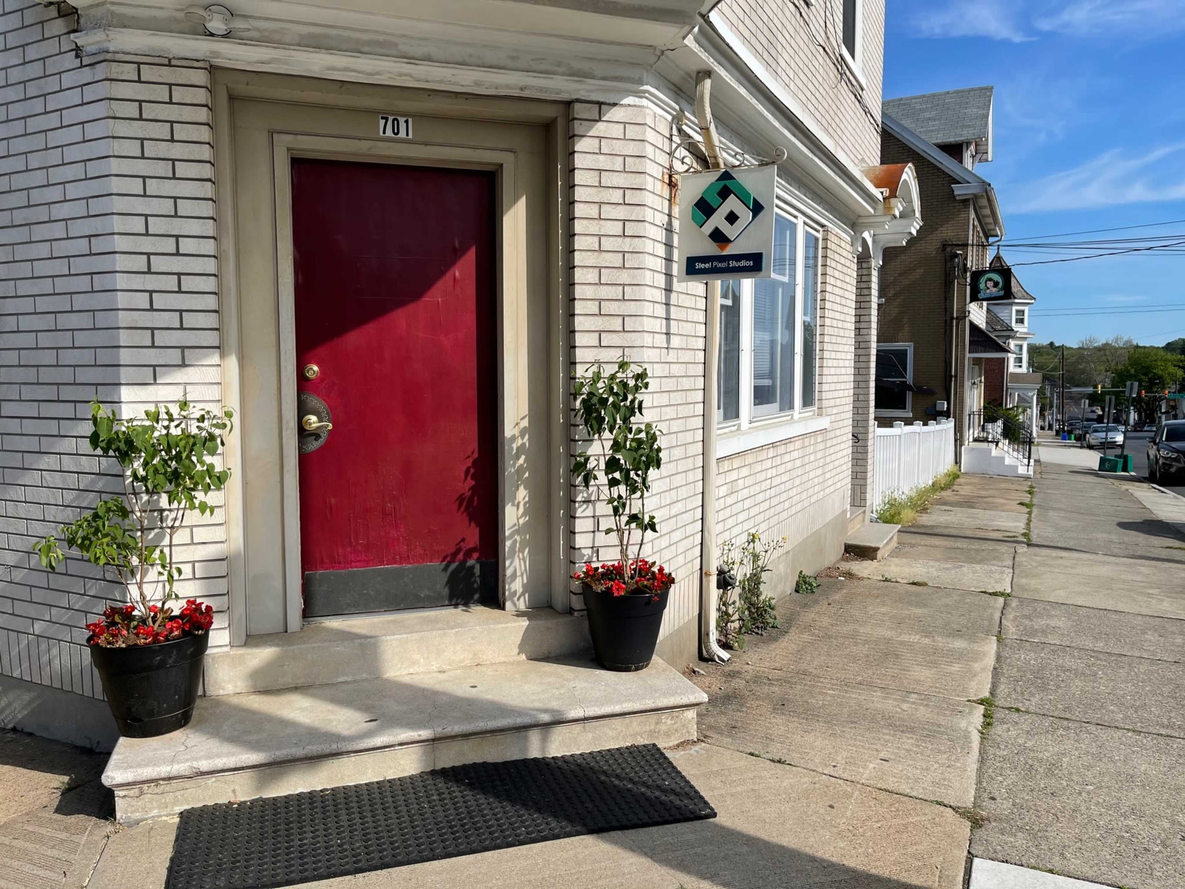 A building entrance with a red door, two potted plants on either side, and a sign near the door, located on a sidewalk next to a street.