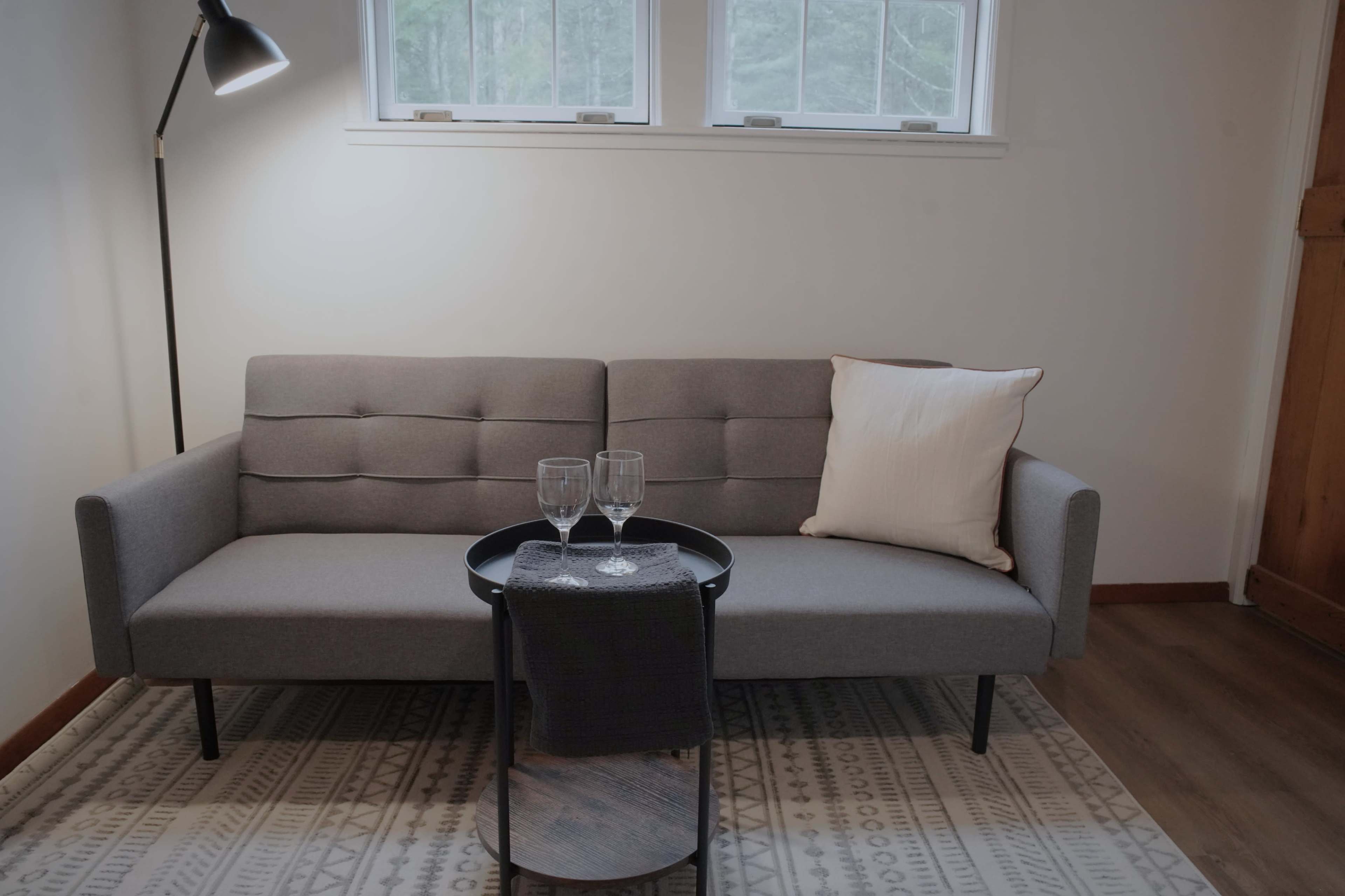 A gray sofa with a cushion sits beside a small table holding two glasses, positioned on a patterned rug in a well-lit room.