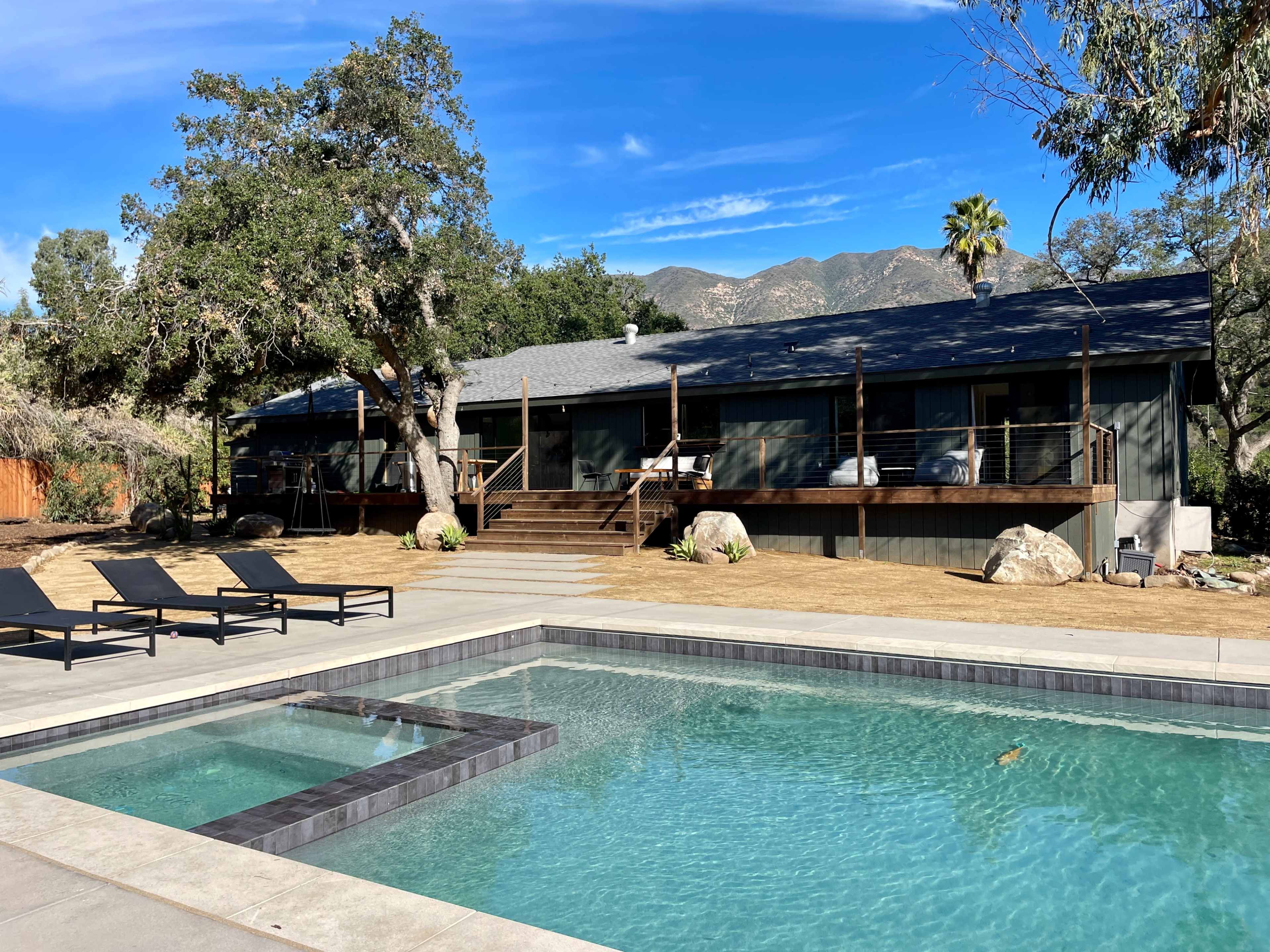 A modern house with a swimming pool is situated in a landscaped outdoor area surrounded by trees and mountains.