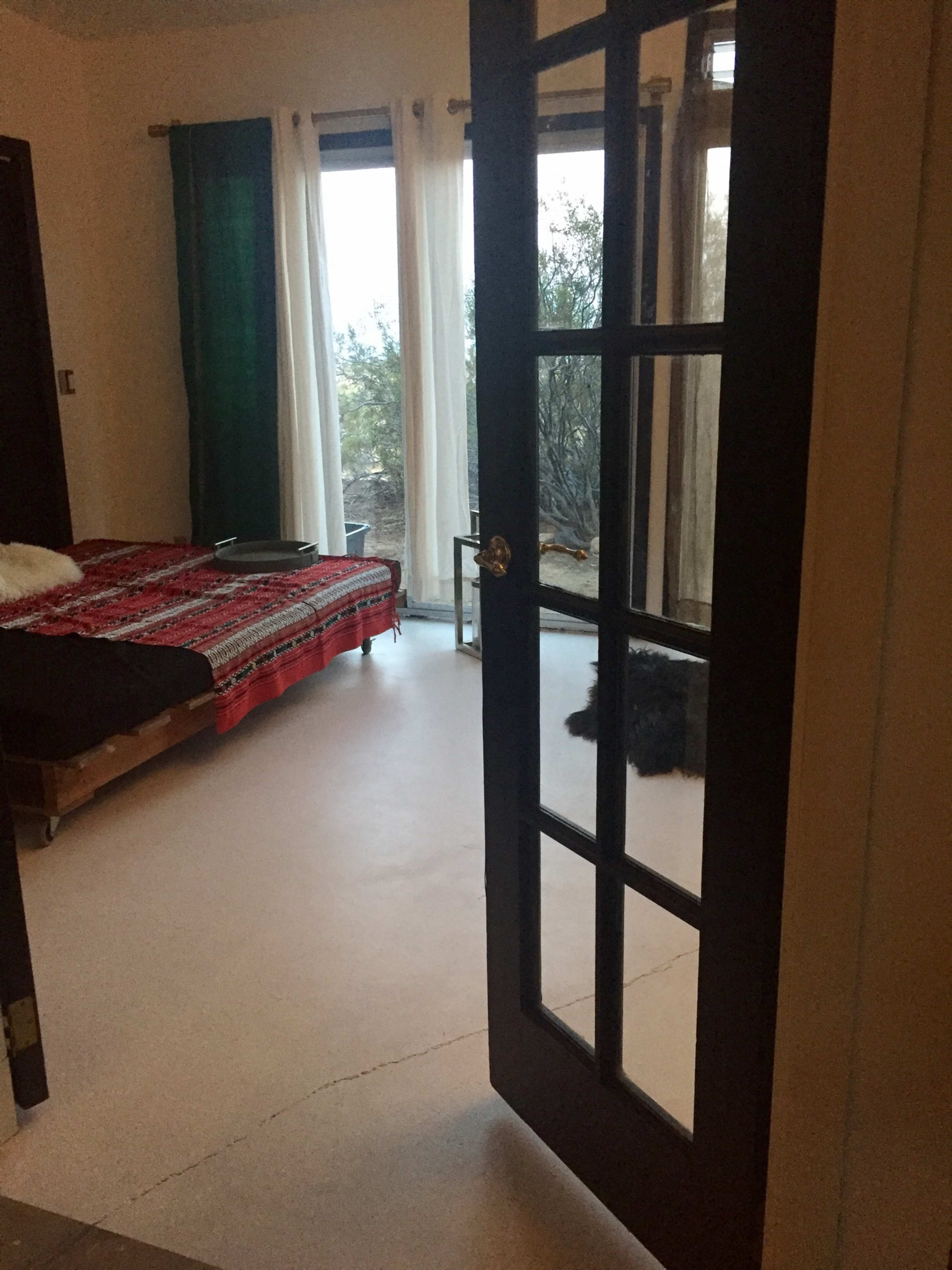 A partially open black door reveals a simple bedroom with a bed covered in a red blanket and a dog lying on the floor near a large window.
