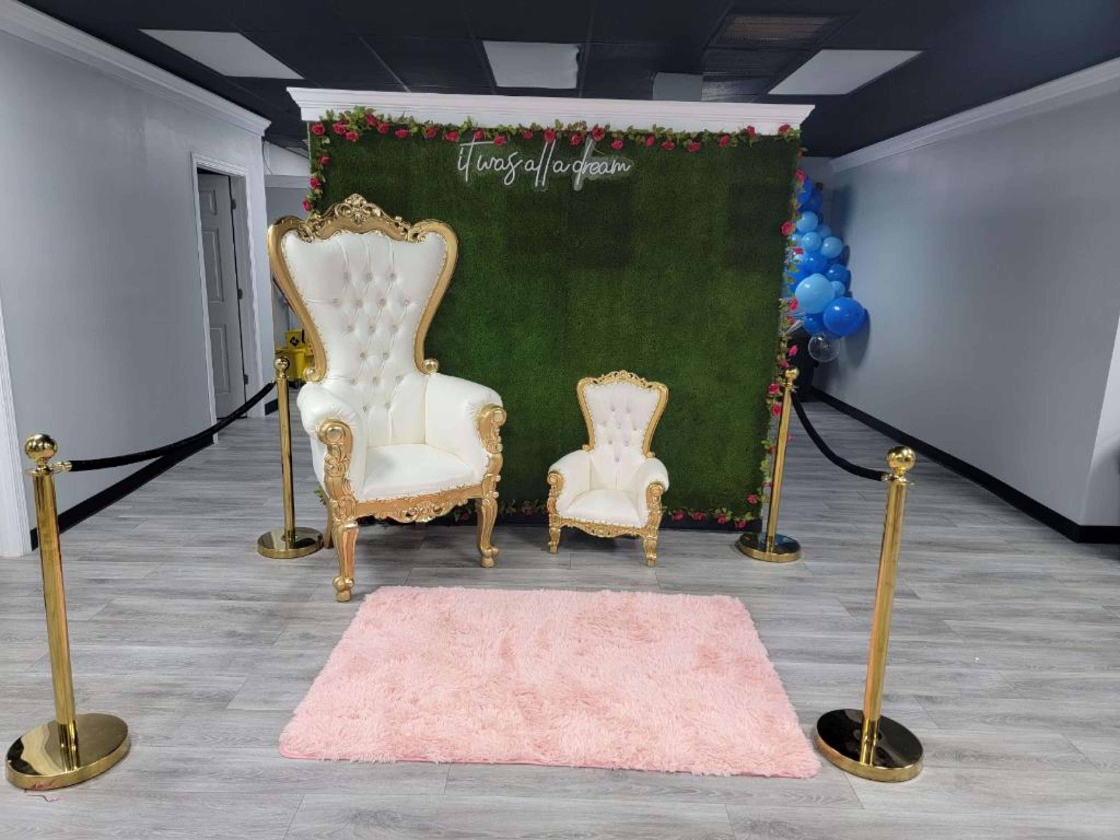 A decorated area featuring a large ornate white chair and a smaller matching chair in front of a green wall with roses, alongside a pink rug and stanchions.