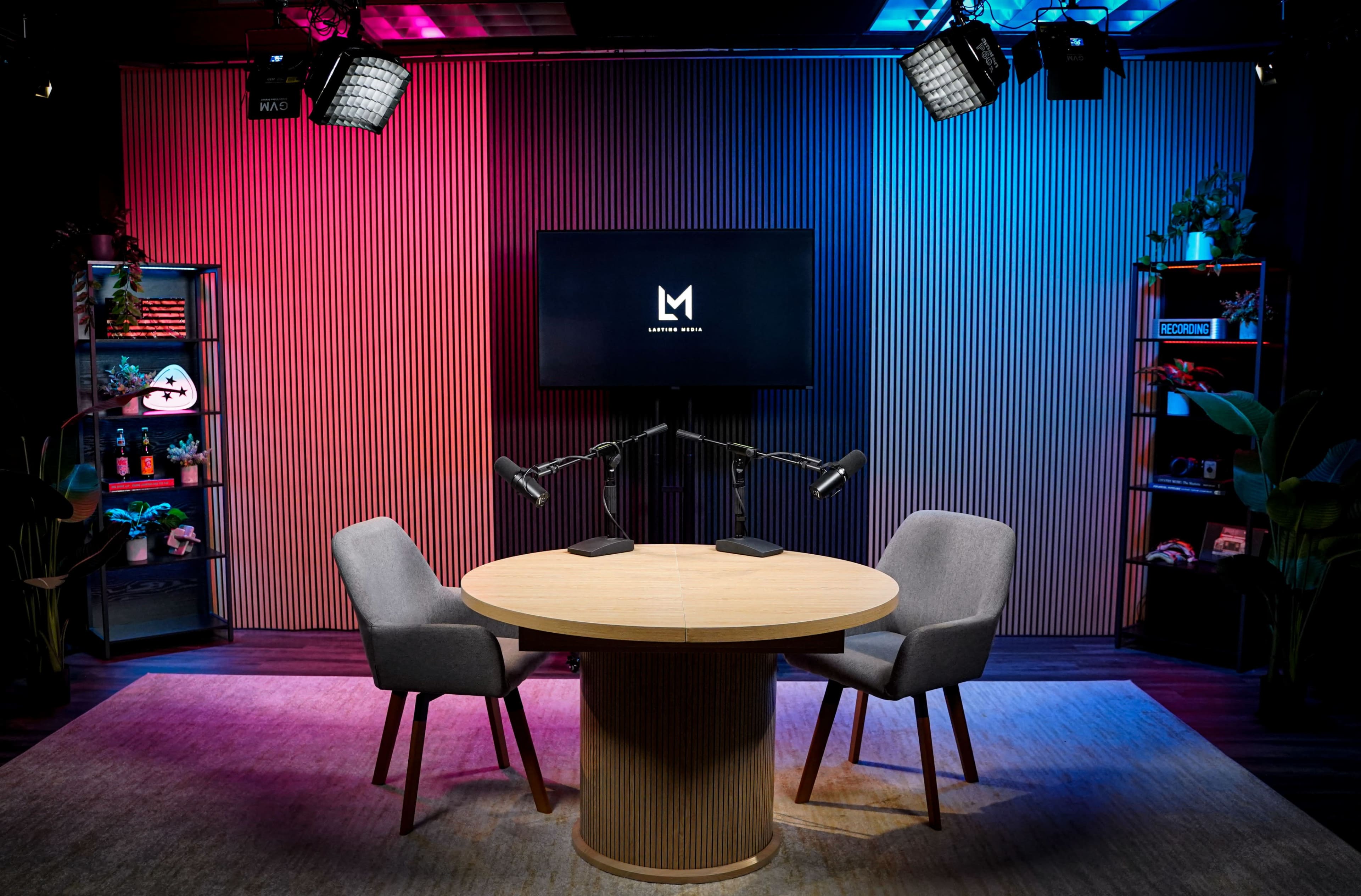 The image shows a modern podcast studio setup with a circular wooden table, two chairs, and a large screen displaying a logo in the background, flanked by colorful lighting and shelves with decorative items.