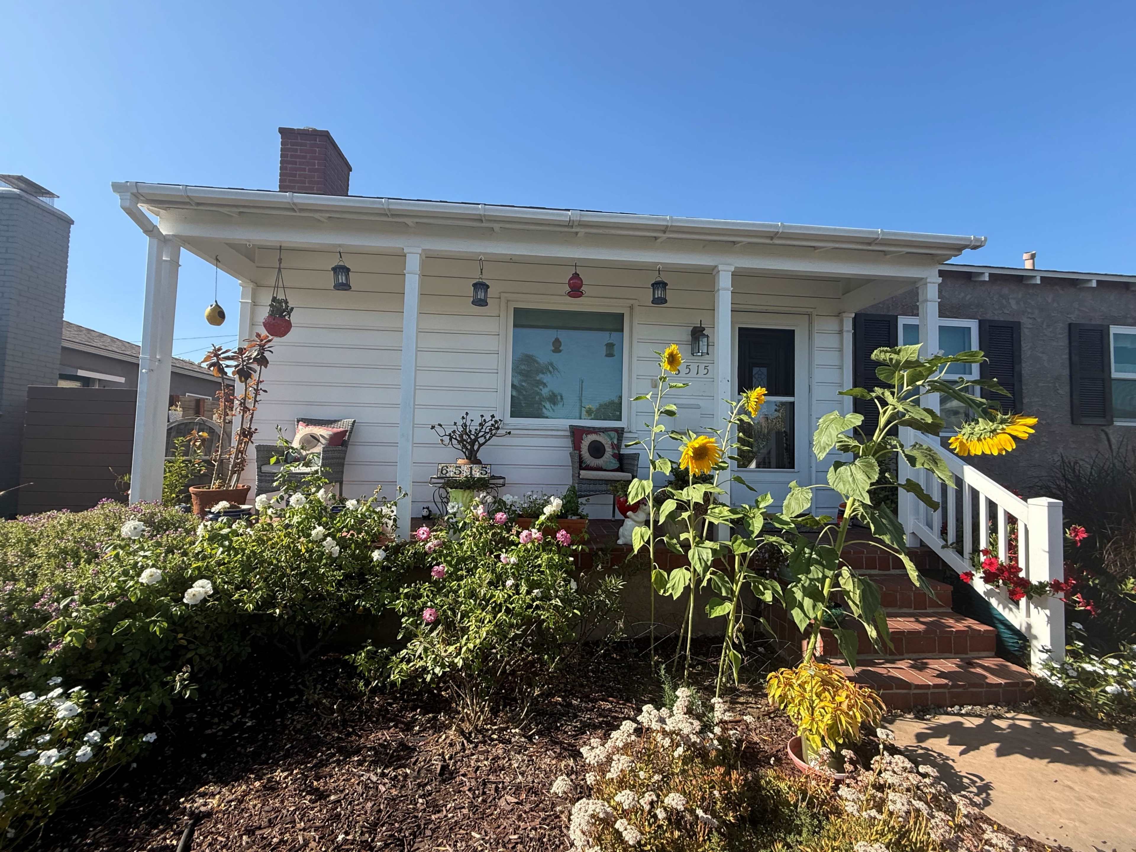 A small white house with a front porch is surrounded by colorful flowers and tall sunflowers under a clear blue sky.