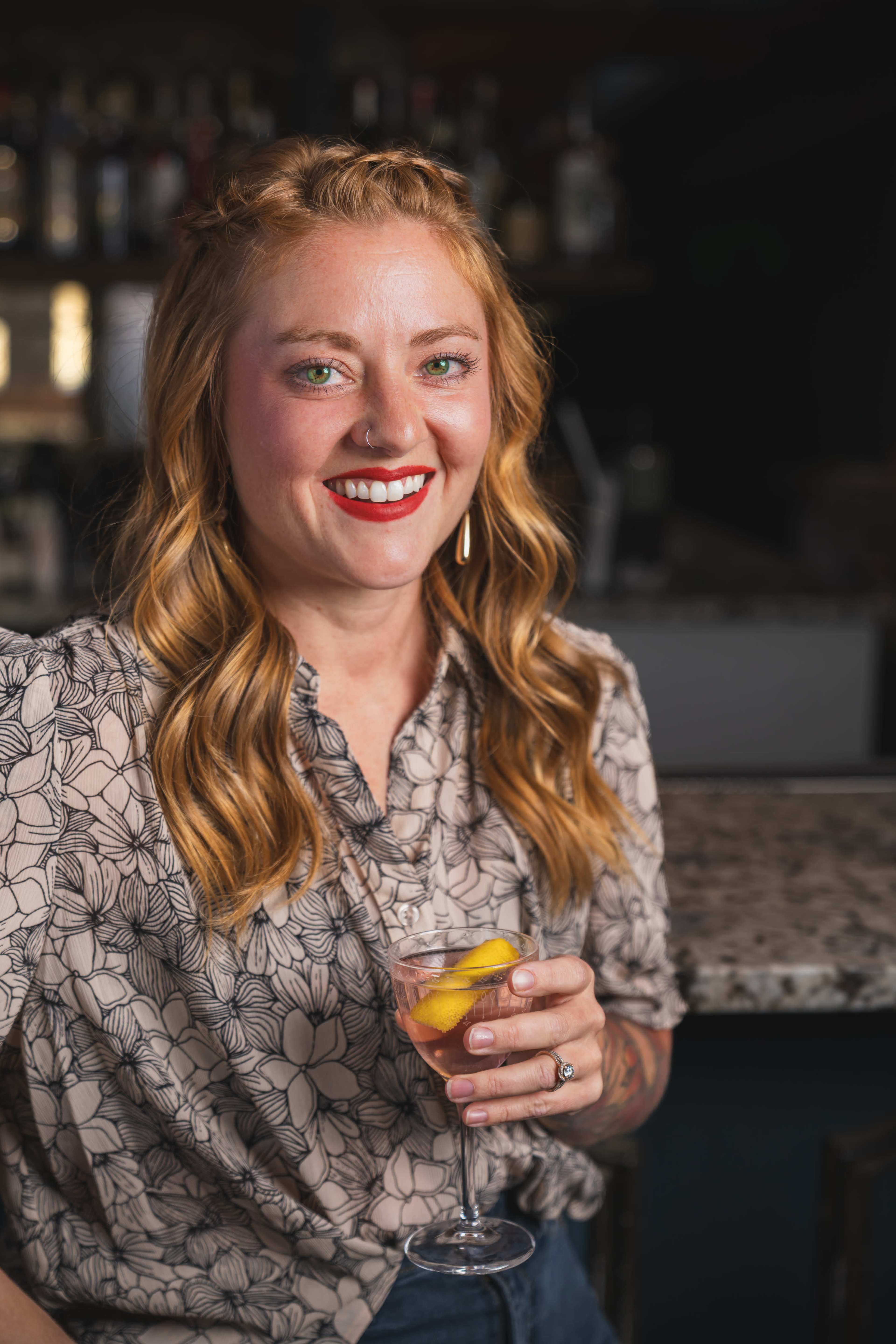 A woman with wavy hair holds a cocktail with a lemon slice while sitting at a bar.