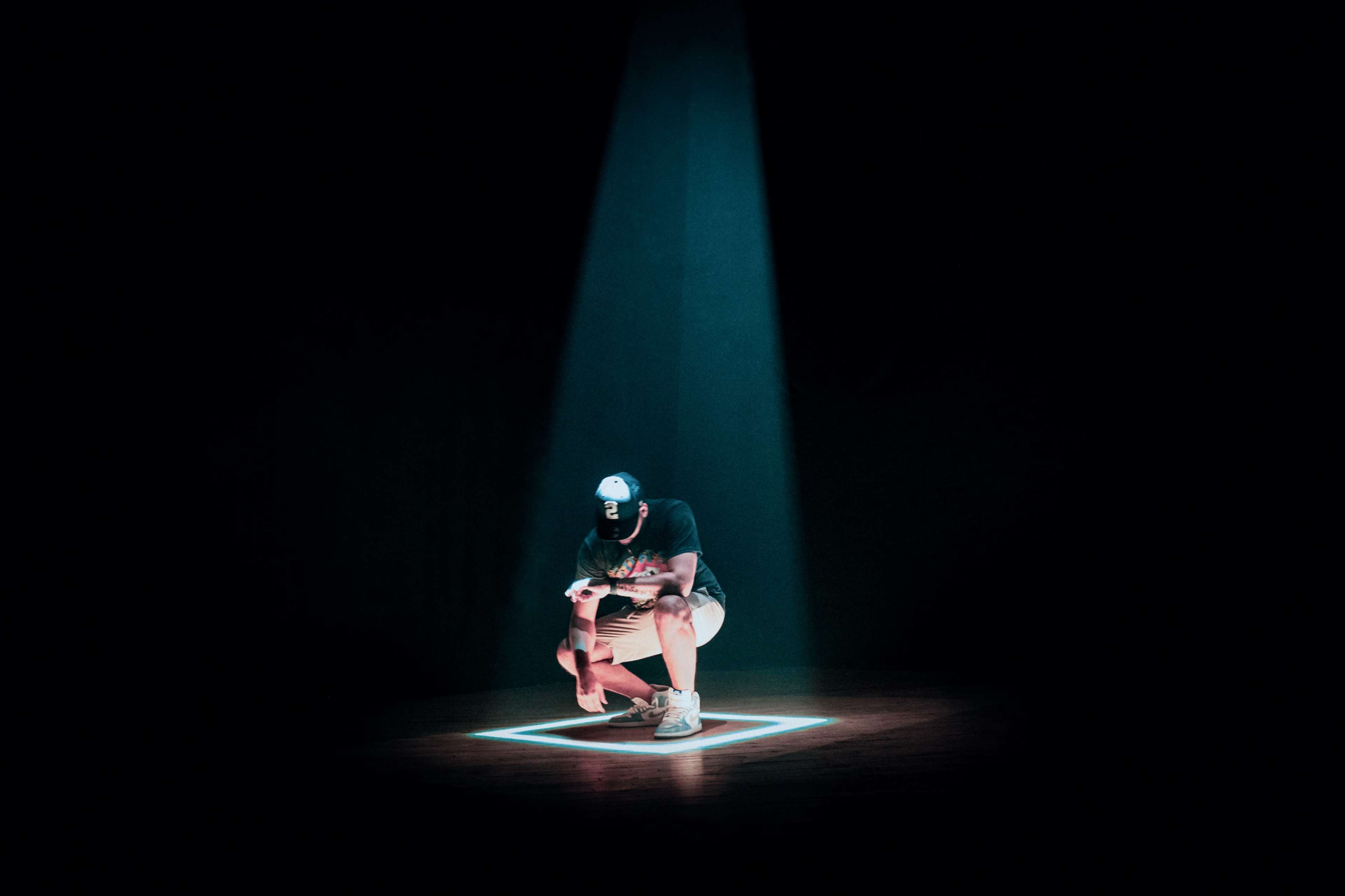 A person squats within a illuminated square on a dark stage, with a beam of light shining down from above.