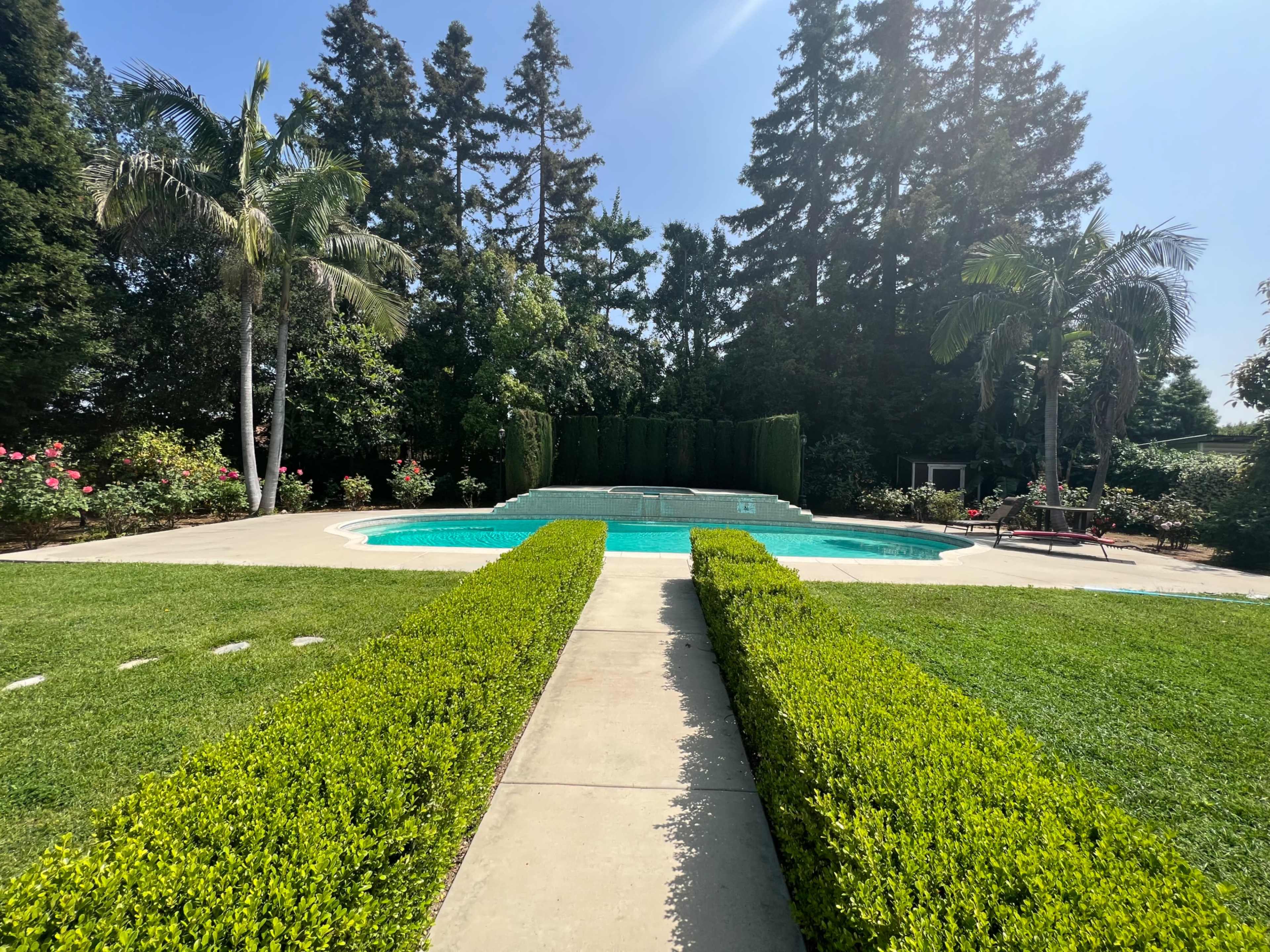 A pool is surrounded by neatly manicured hedges and a lawn, with tall trees and landscaped flower beds in the background.