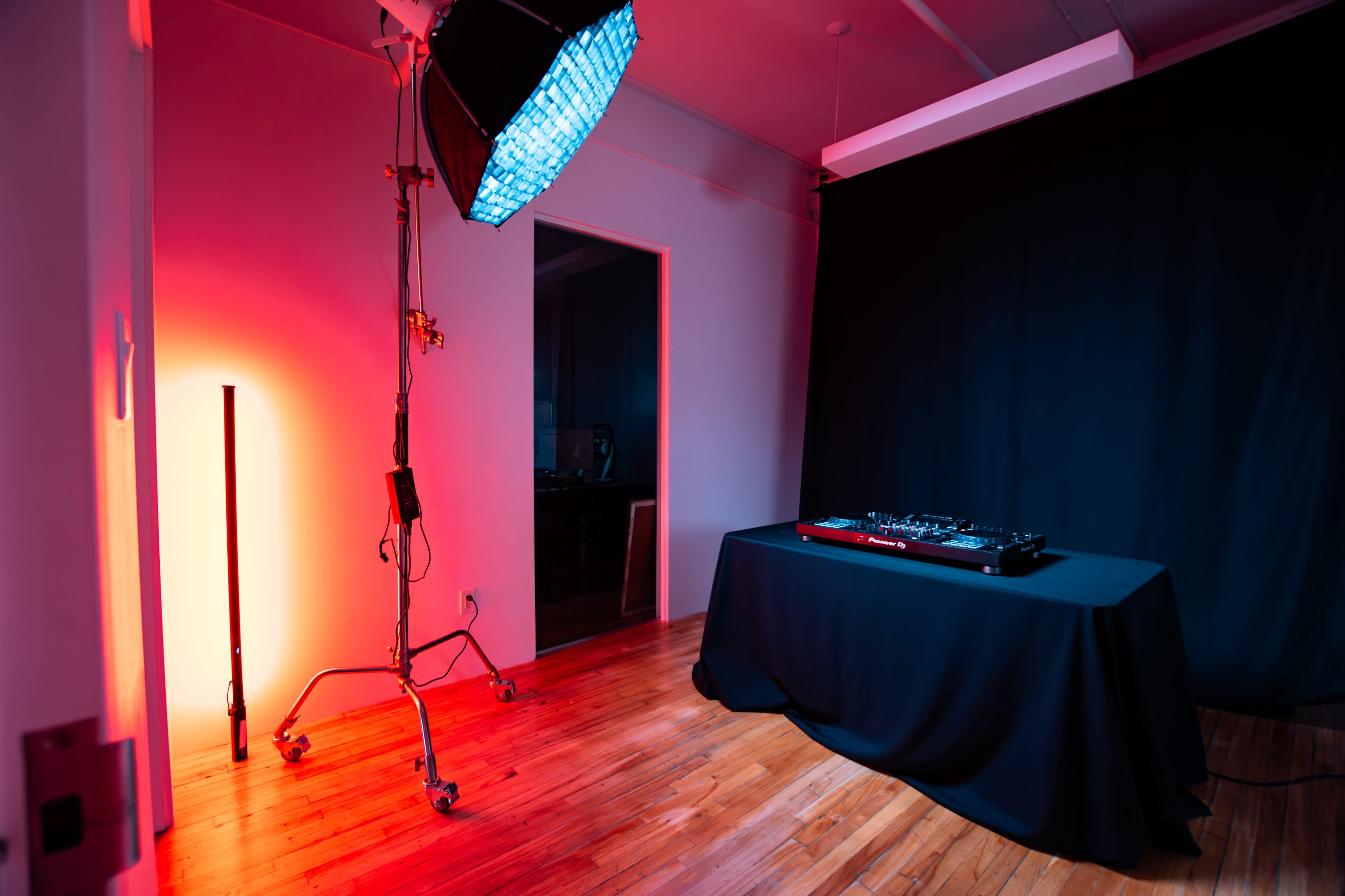 The image shows a dimly lit studio space featuring a black table with a DJ controller, illuminated by colorful lighting and a black backdrop.