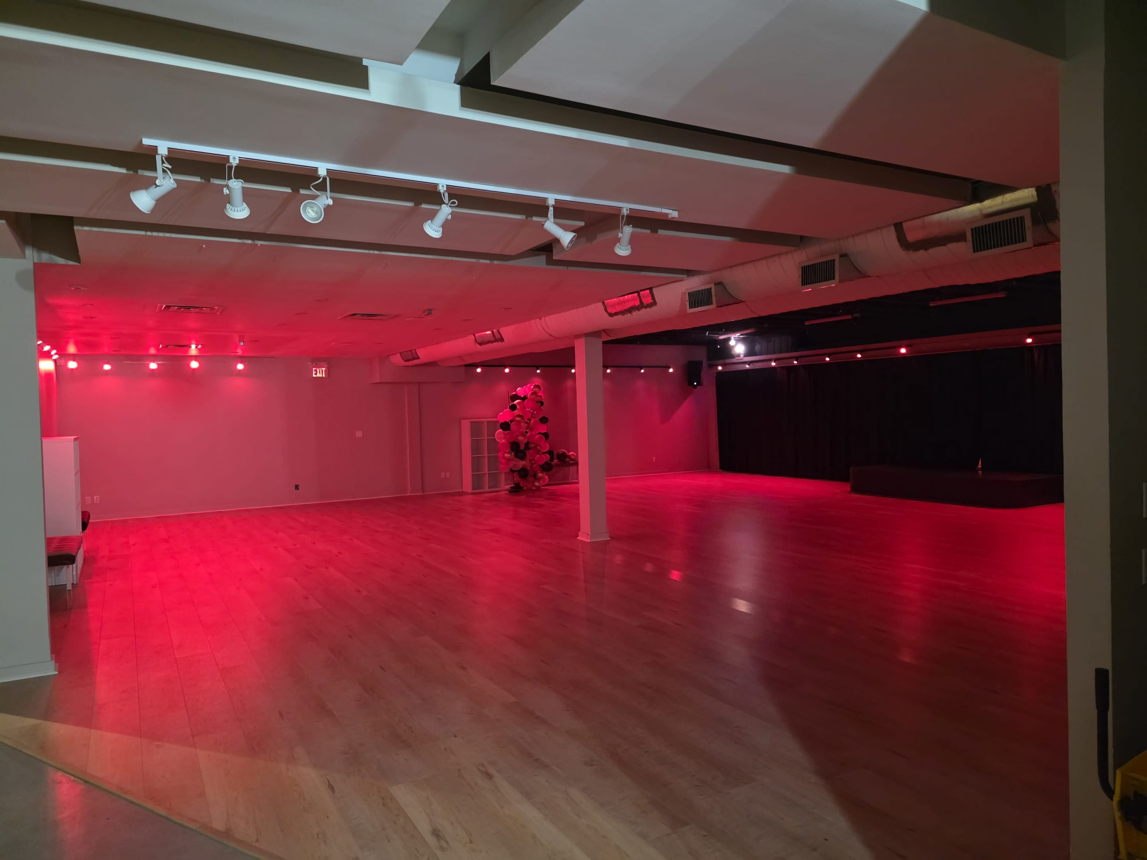 The image shows an empty studio space with hardwood flooring, illuminated by red lighting from above, and a black curtain backdrop on one side.