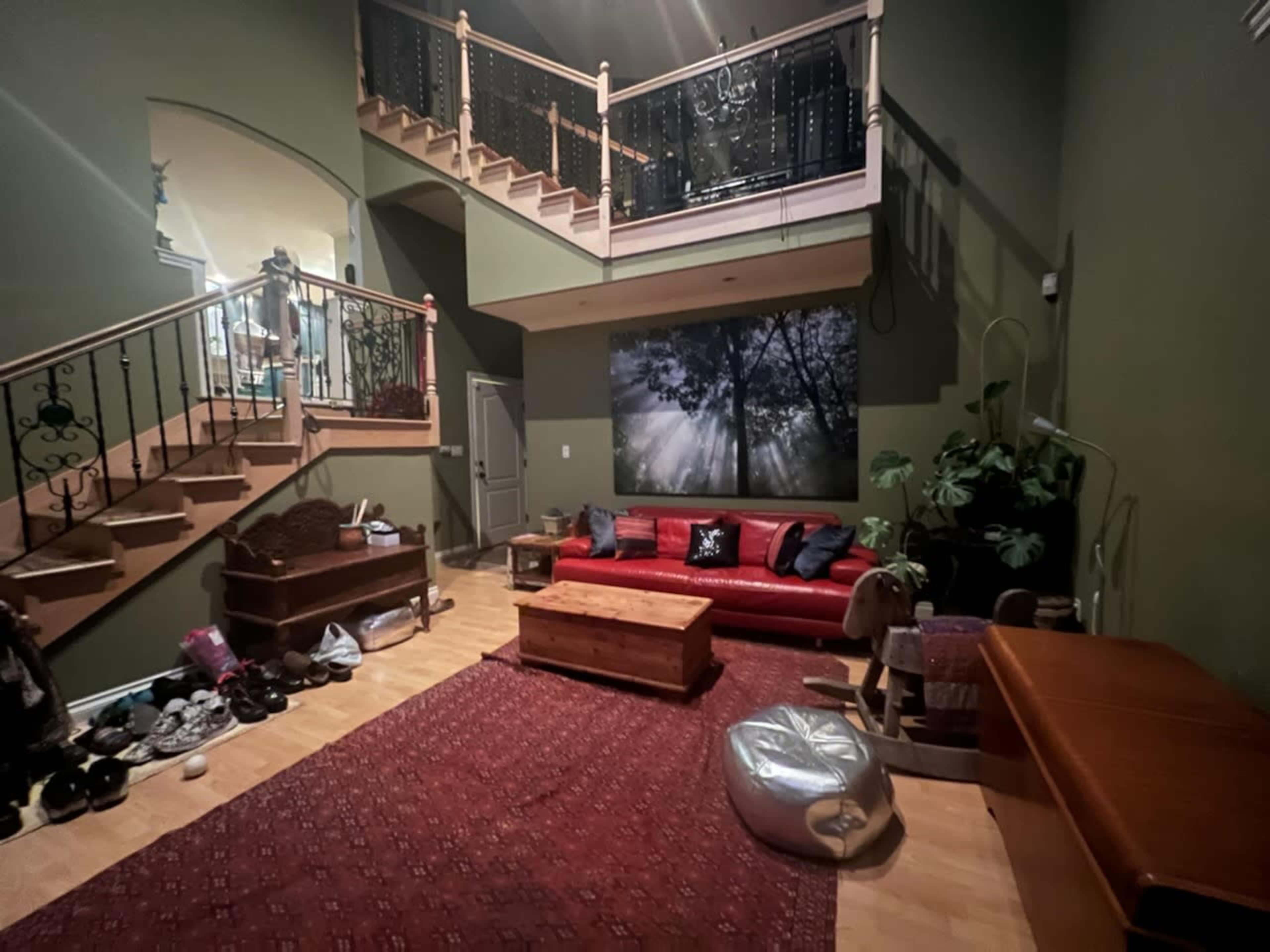 A spacious living room features a red sectional sofa, a wooden coffee table, and a large staircase leading to an upper level, with a wall adorned by a tree-themed mural.