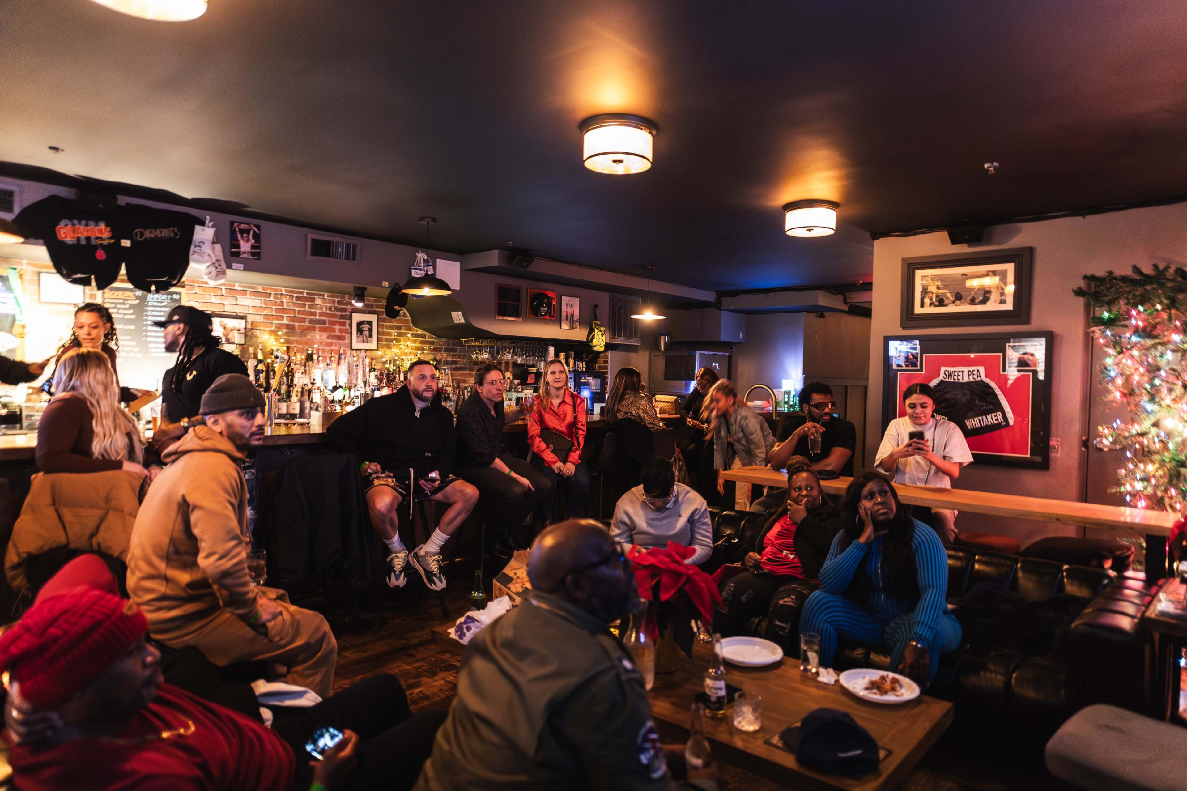 A diverse group of people is seated in a cozy bar, watching a performance or event on a screen while food and drinks are placed on tables.