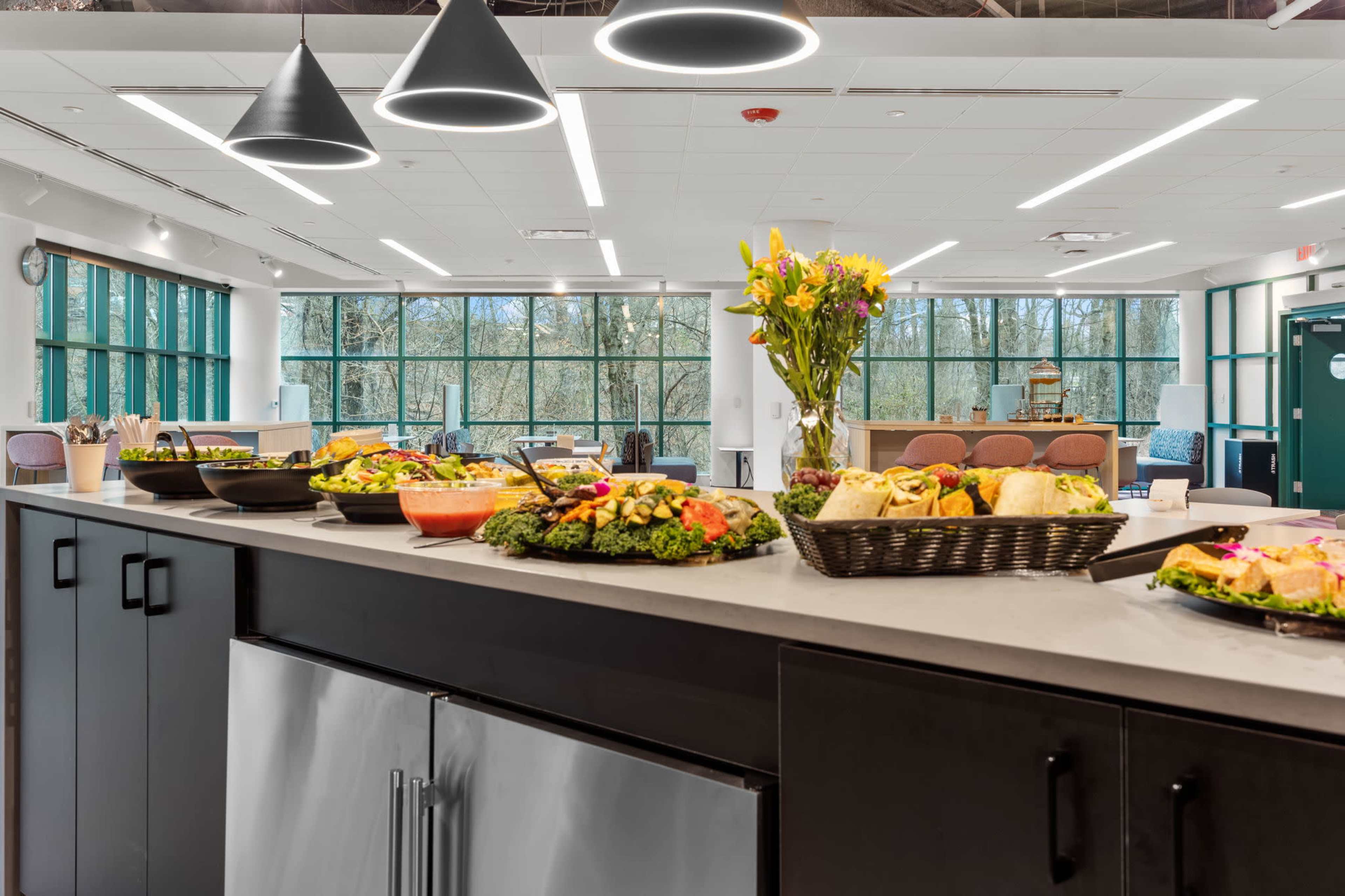 A buffet table displays an assortment of colorful dishes, with large windows revealing a view of trees in the background.