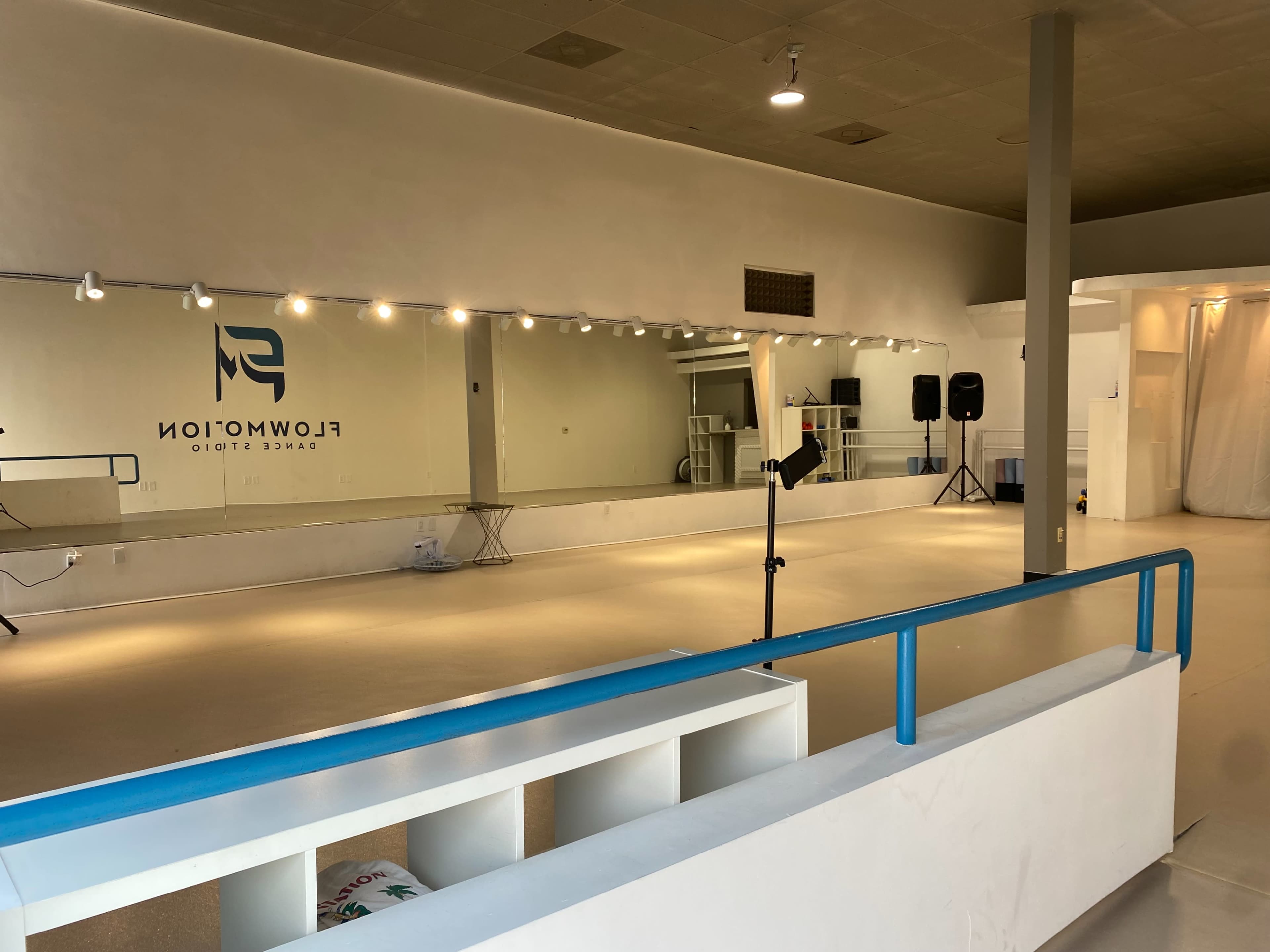 A spacious dance studio with a large mirrored wall, light fixtures above, and a blue railing separating the viewing area from the dance floor.