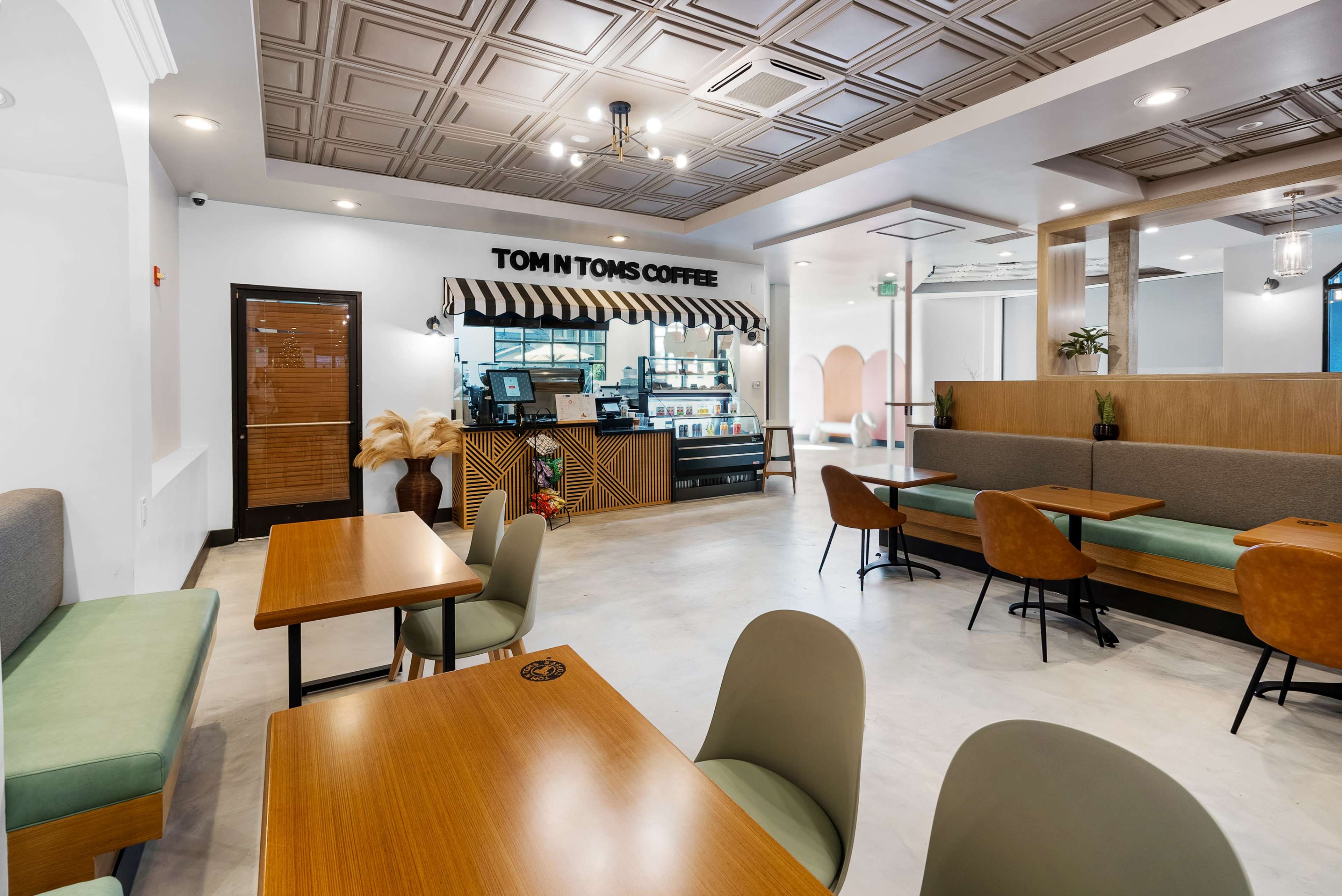 The interior of a coffee shop features wooden tables, green and brown seating, and a service counter with a striped awning.