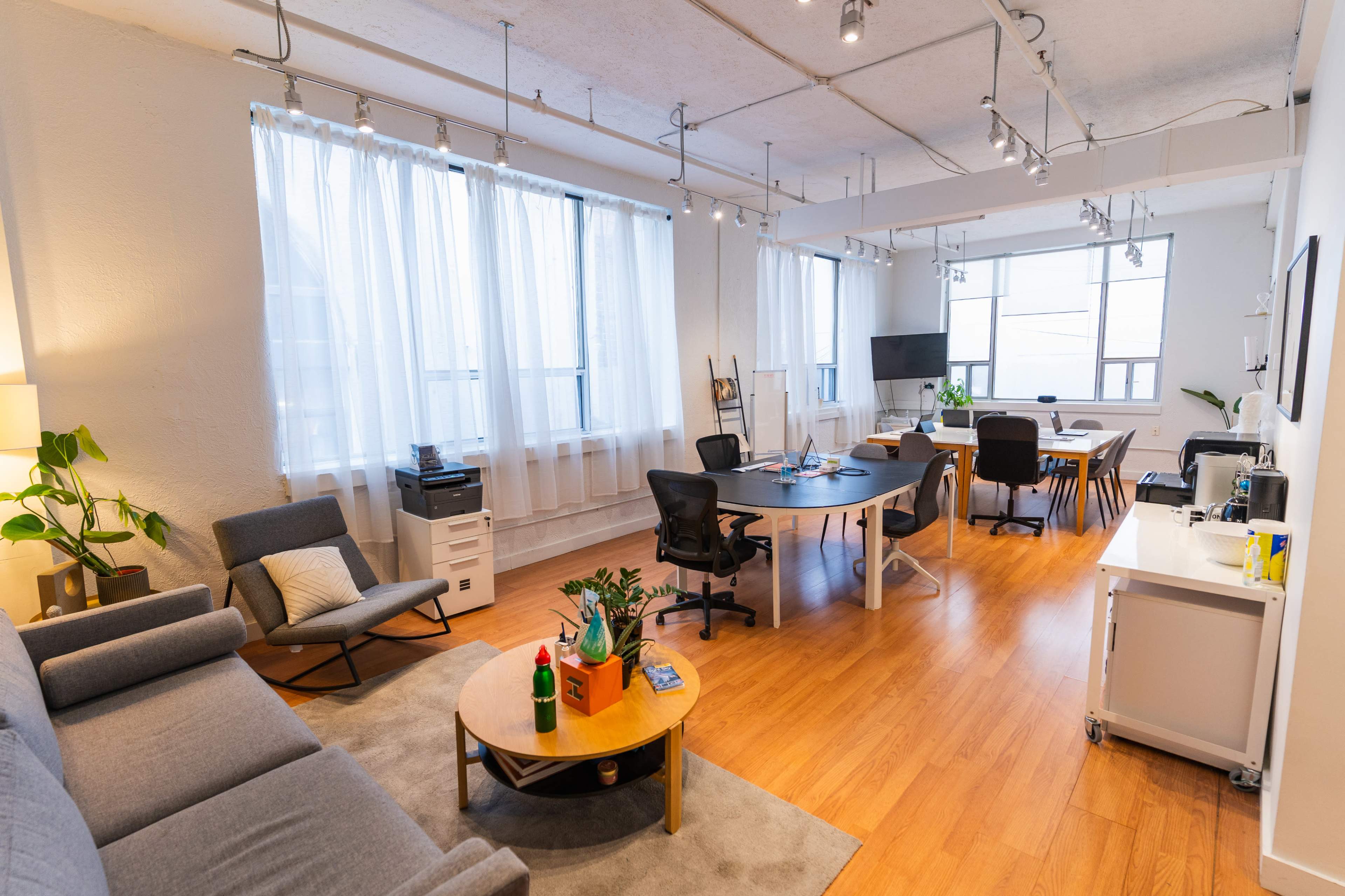 The image shows a bright, open office space with a seating area, a large table for meetings, and several plants.