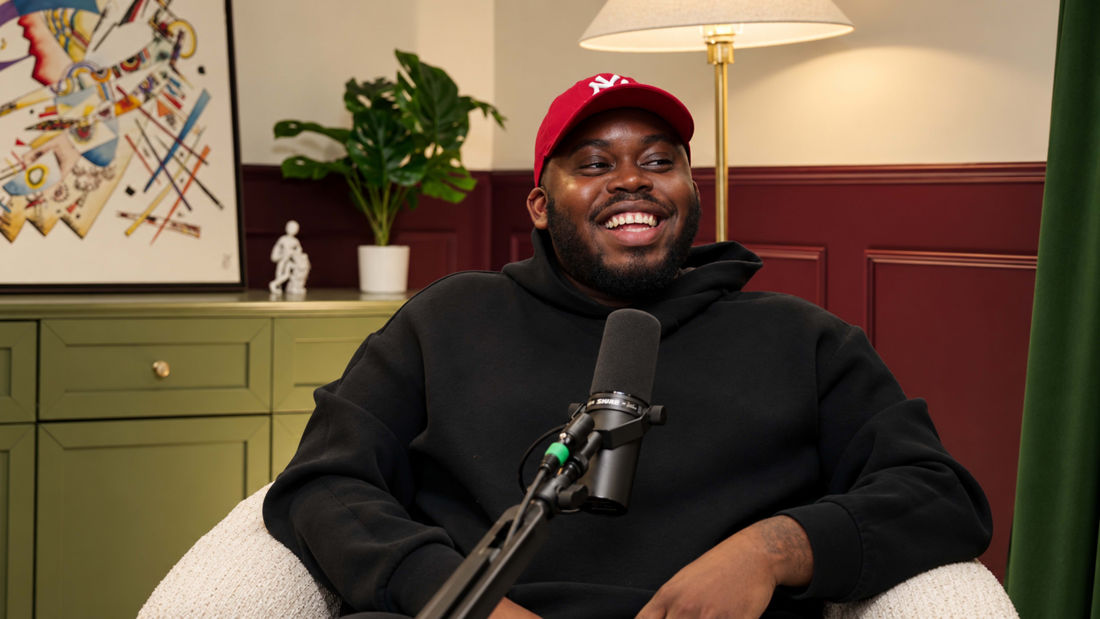 A man wearing a black hoodie and a red cap sits comfortably in a chair, smiling while a microphone is positioned in front of him.