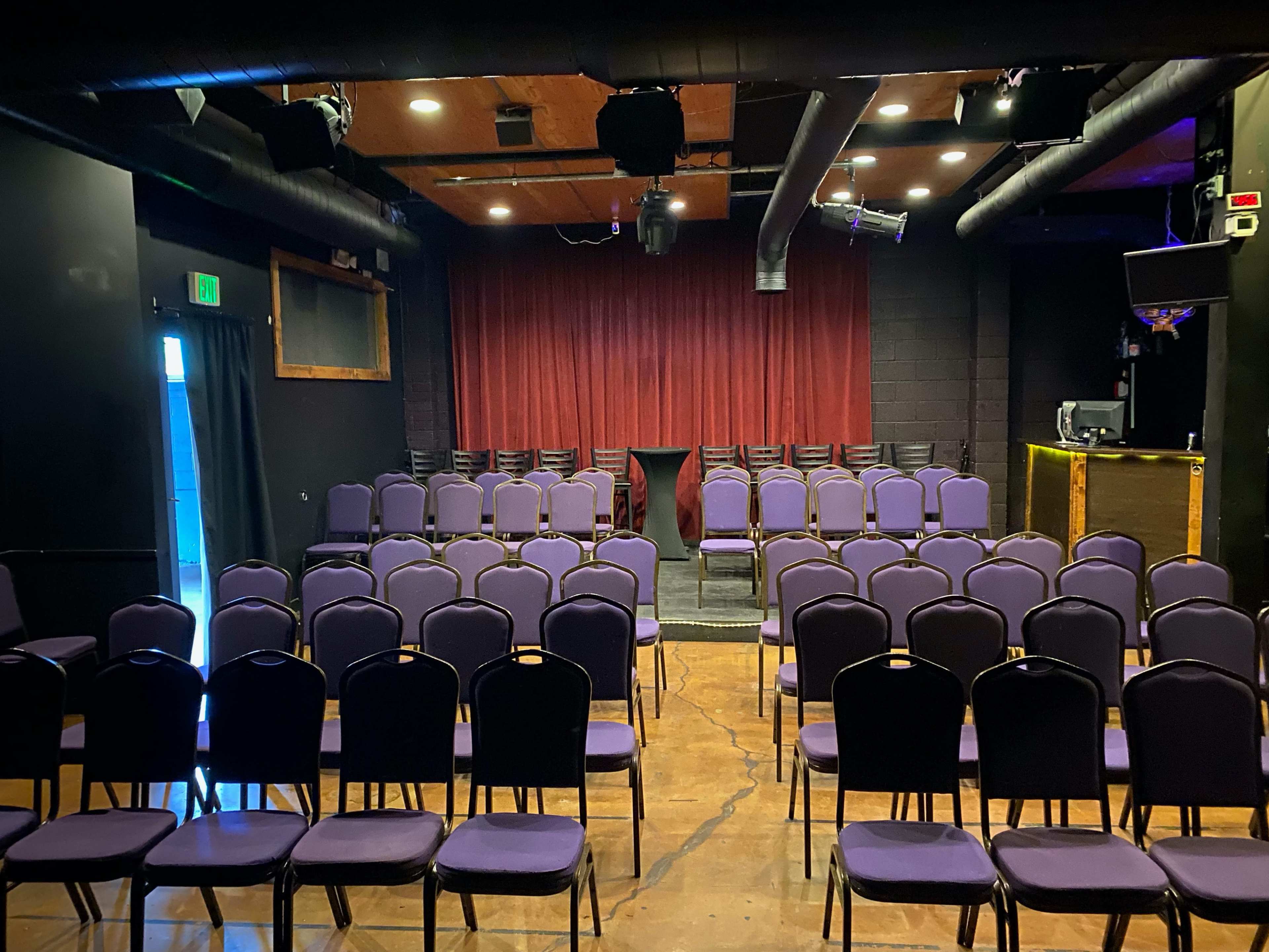 An empty performance space with rows of purple chairs facing a small stage draped in red curtains.