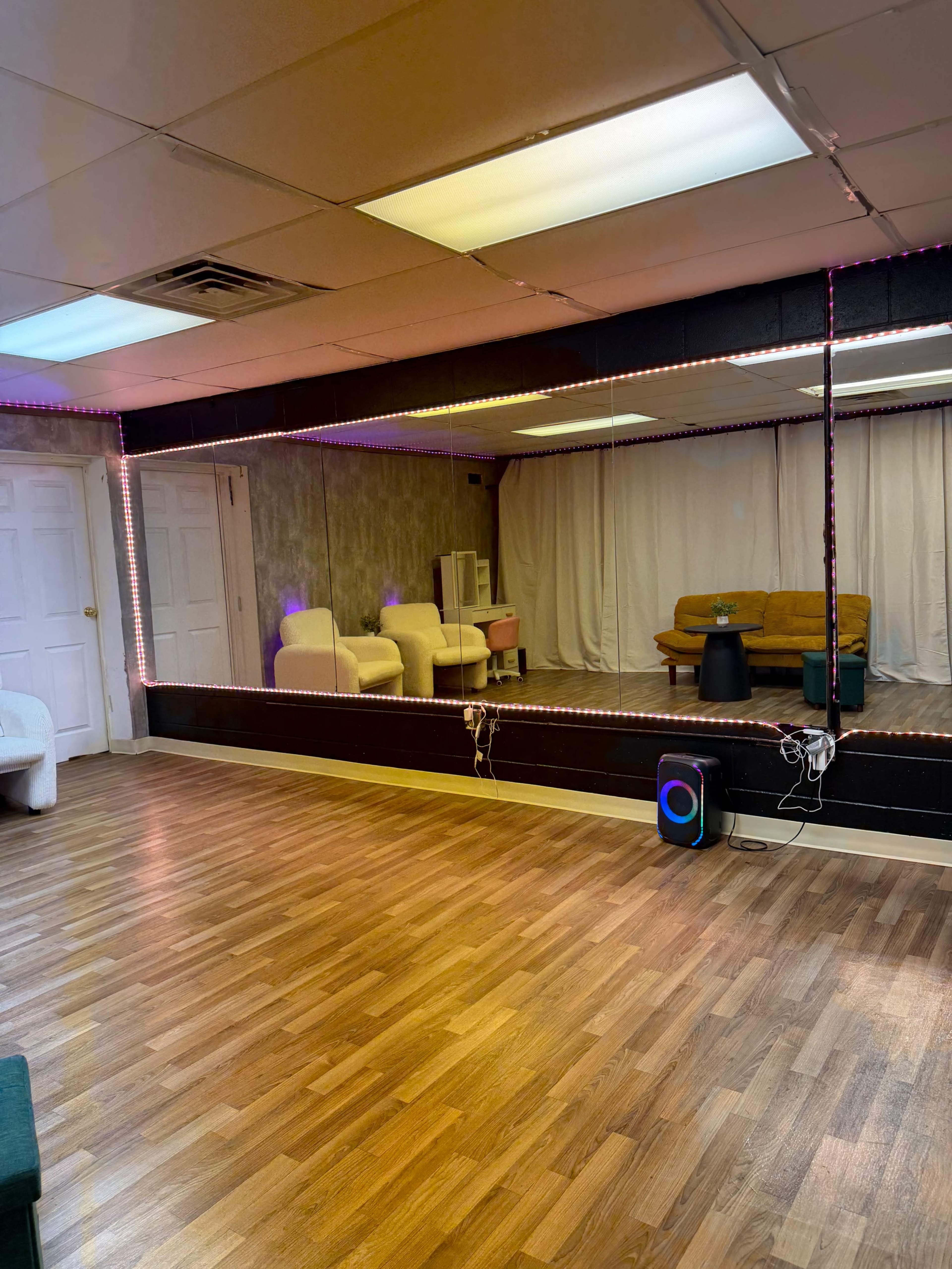 The image shows a spacious, well-lit room with wooden flooring, a large mirror, a couch in the background, and decorative lighting along the edges.