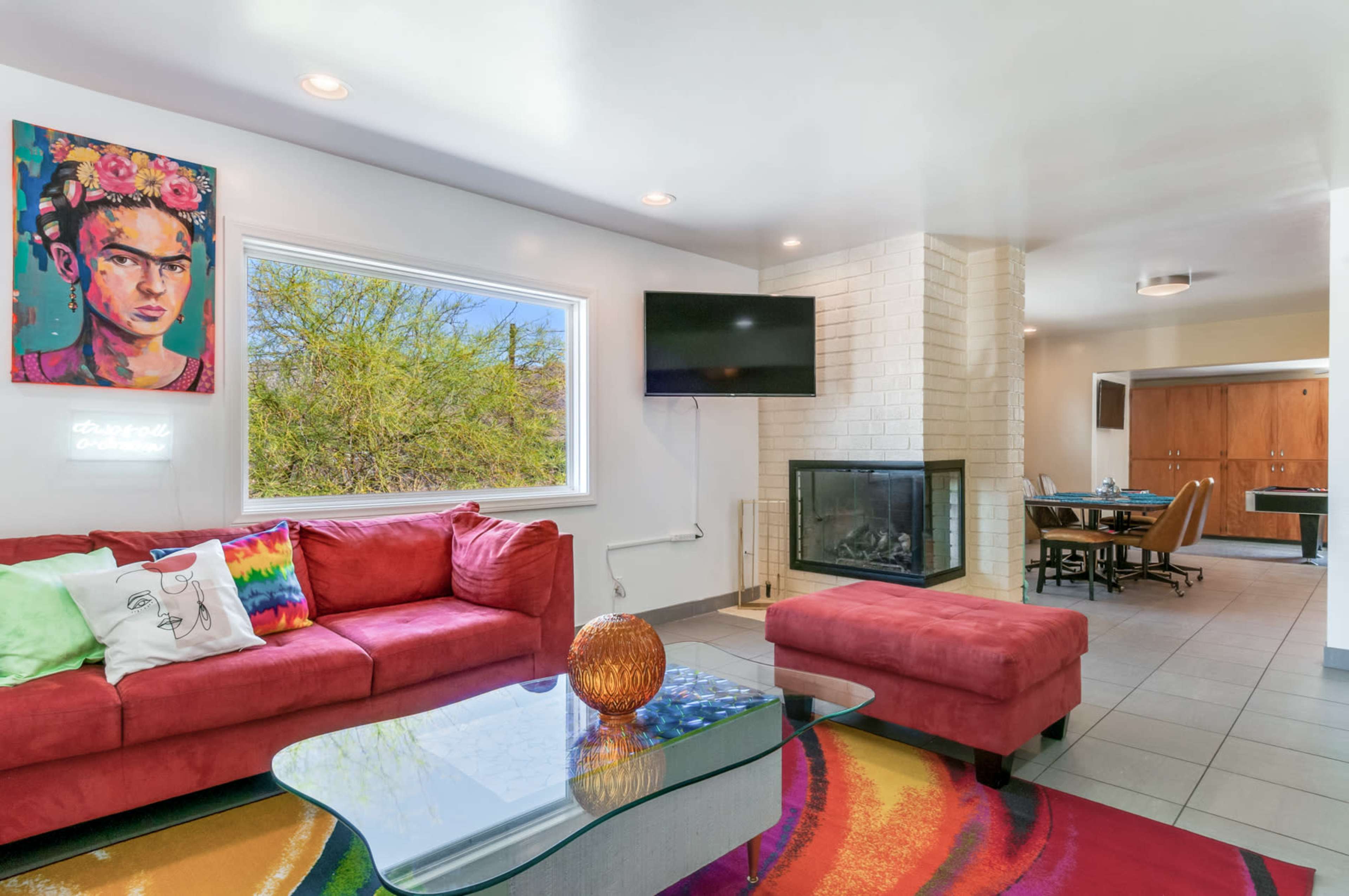 The living area features a red sectional sofa, a glass coffee table, and a wall-mounted television, with a fireplace and dining area visible in the background.