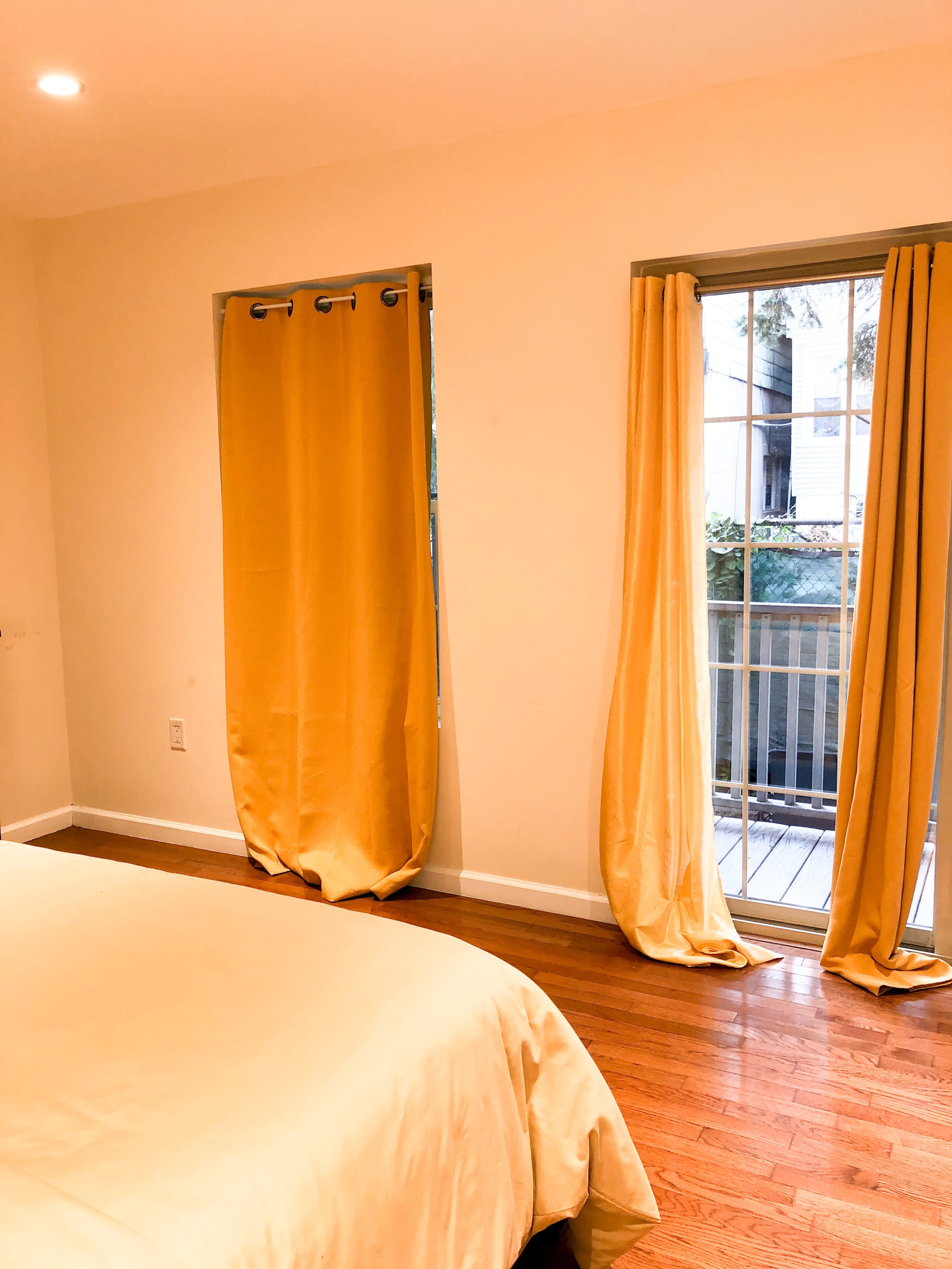 A bedroom features a bed with a light-colored duvet and two windows dressed with yellow curtains.