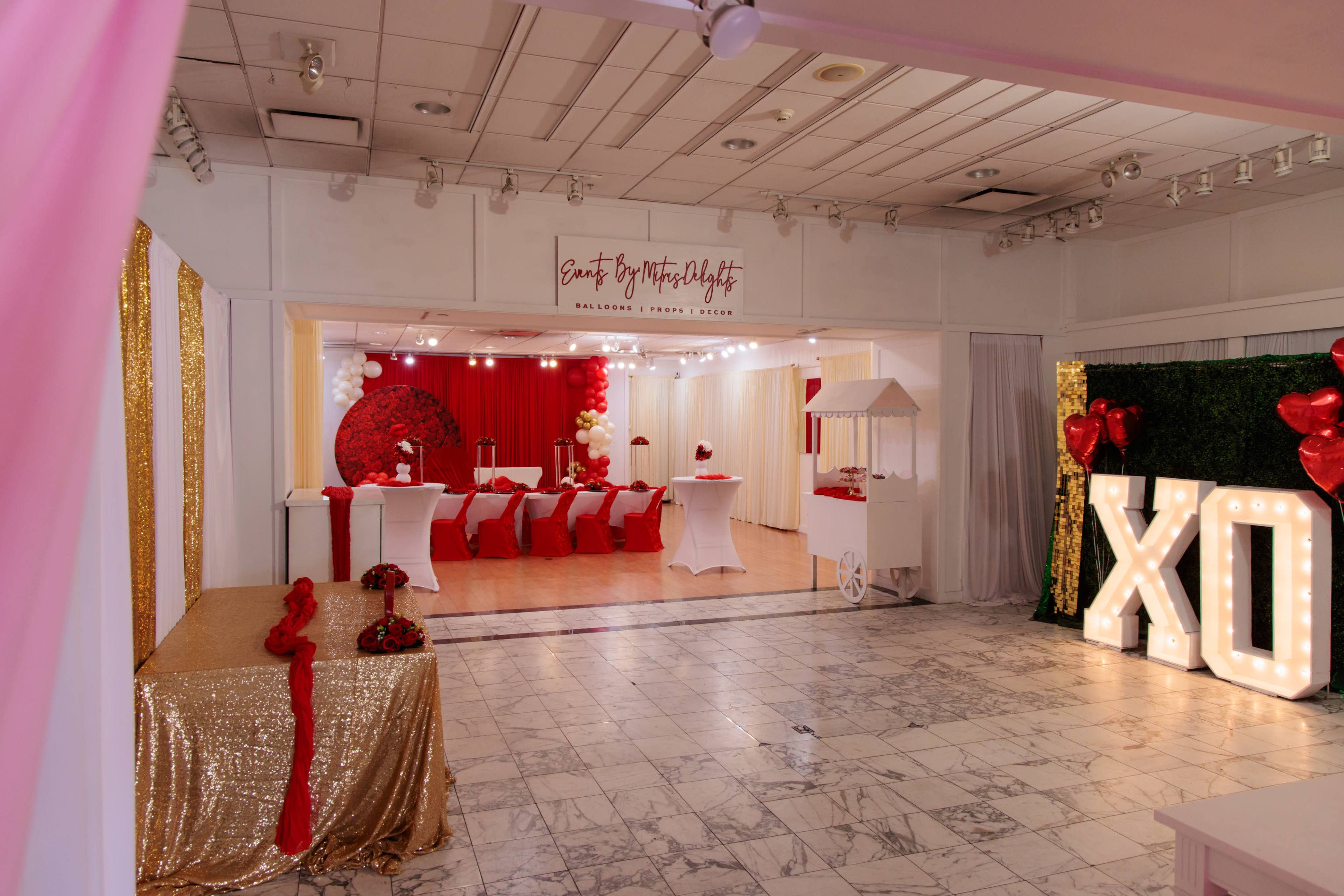 The image shows a decorated event space featuring a red and white theme, with tables set for dining, a dessert station, and large illuminated letters spelling "XO."