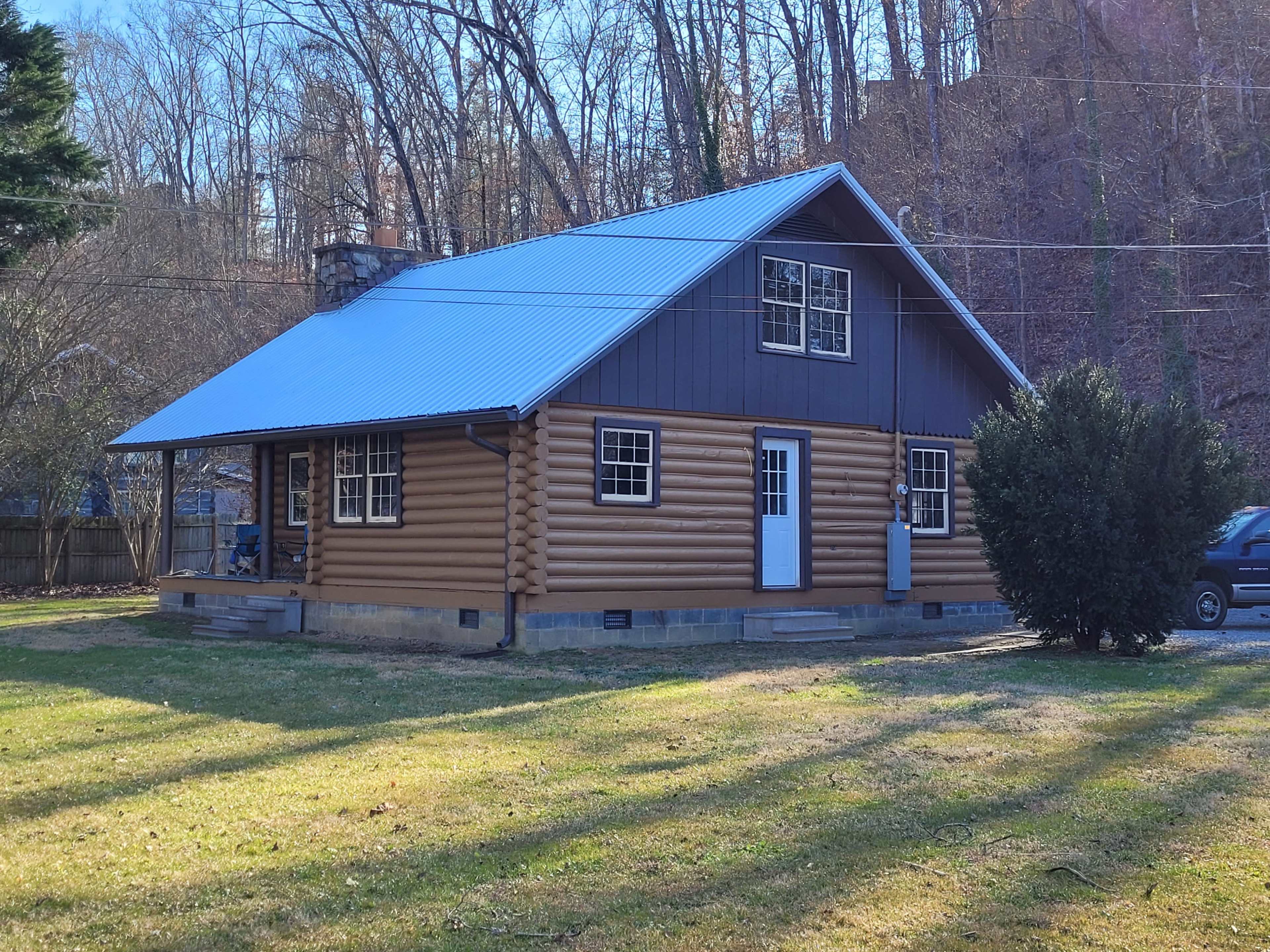 A log cabin with a blue metal roof and white trim is situated on a grassy area surrounded by trees.