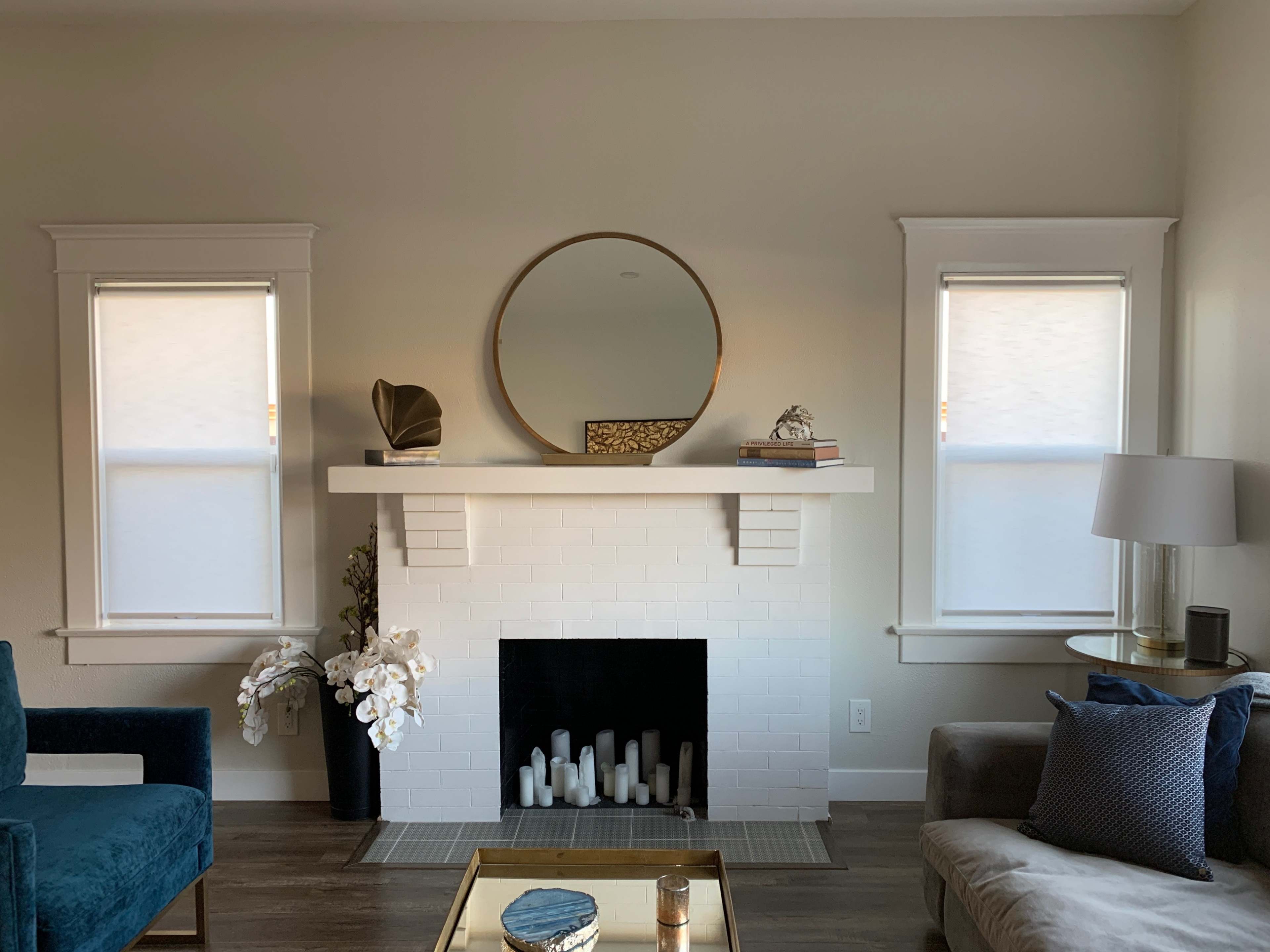 The image shows a living room with a white brick fireplace, a large round mirror above it, and two windows flanking the mantle, decorated with a few decorative items and plants.