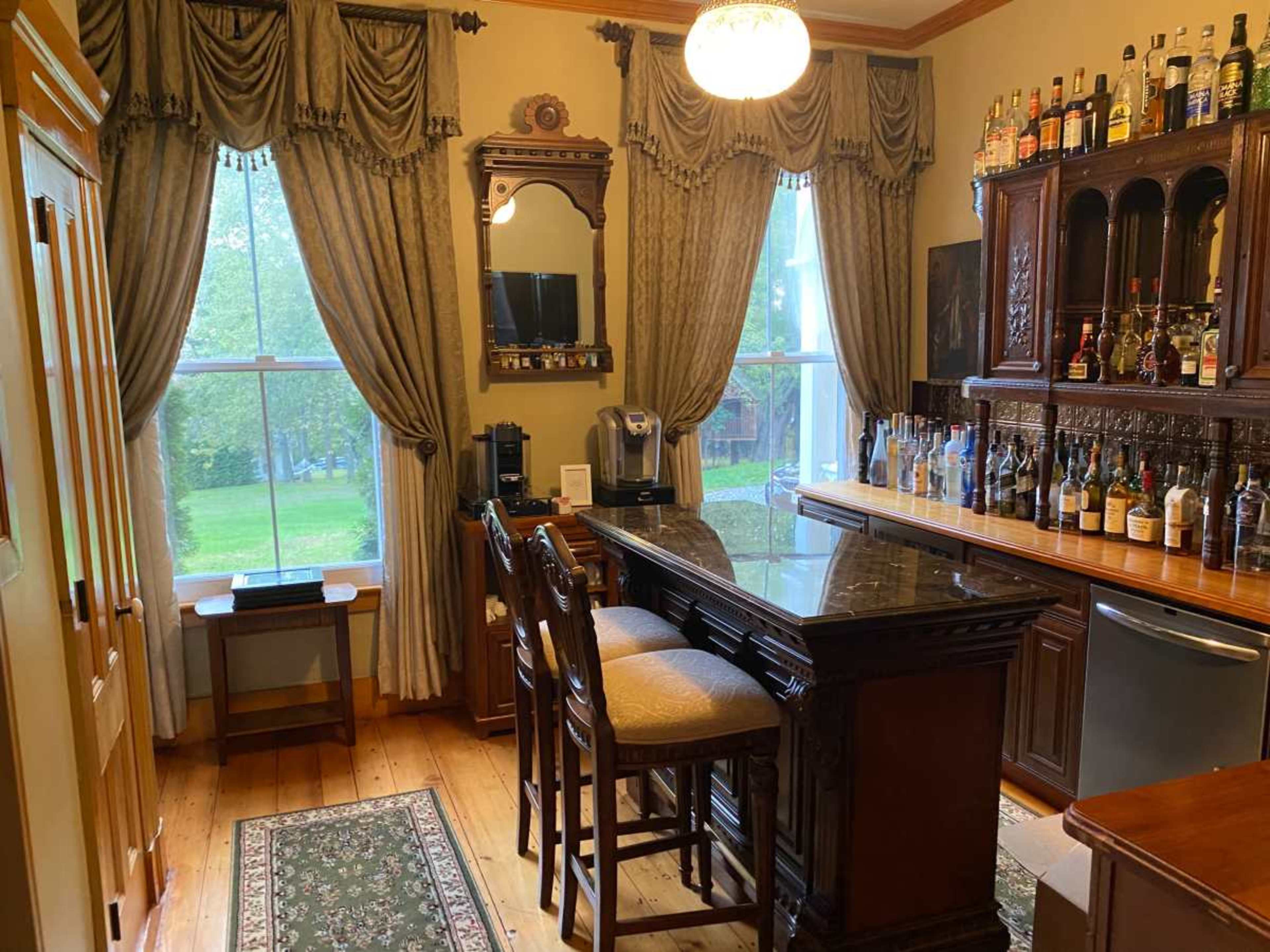 The image shows a wooden bar with high stools, situated in a room featuring large windows dressed with patterned drapes, and a bookshelf containing various bottles and glassware.