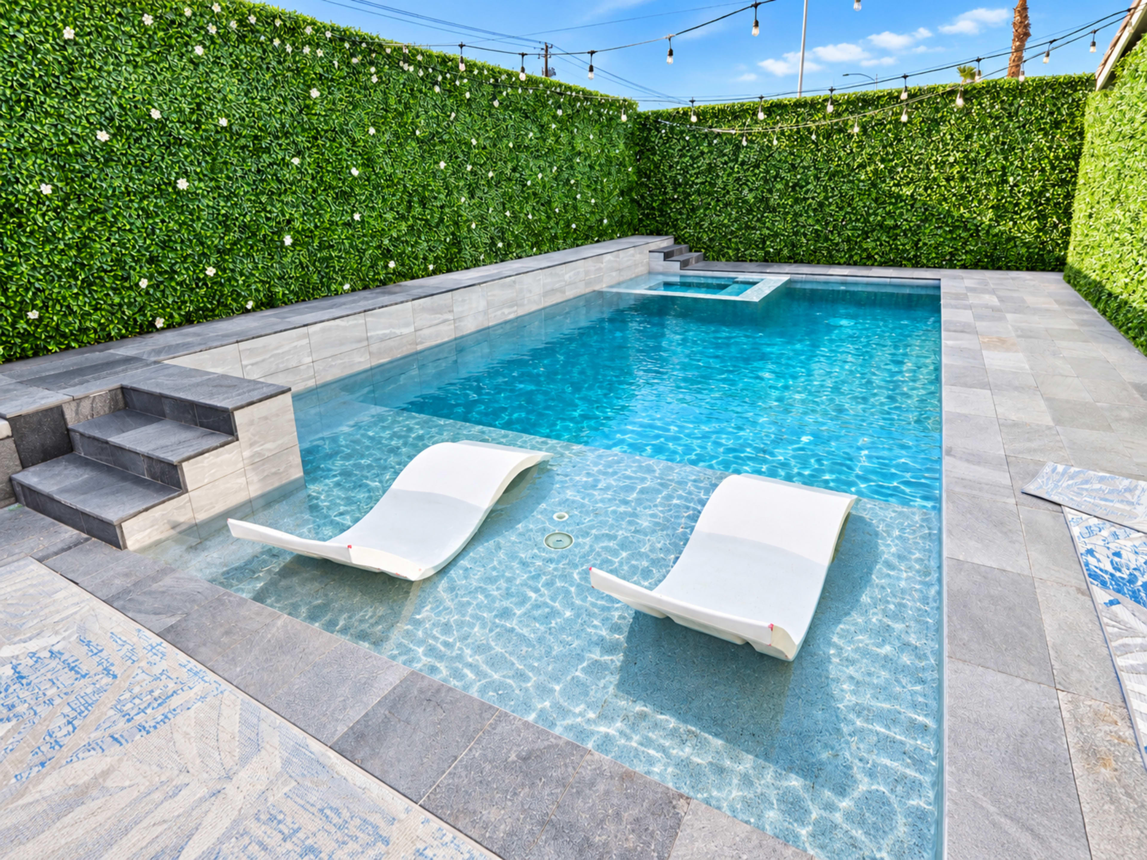 The image shows a modern outdoor swimming pool with two lounge chairs on the edge, surrounded by green hedges and a clear blue sky.