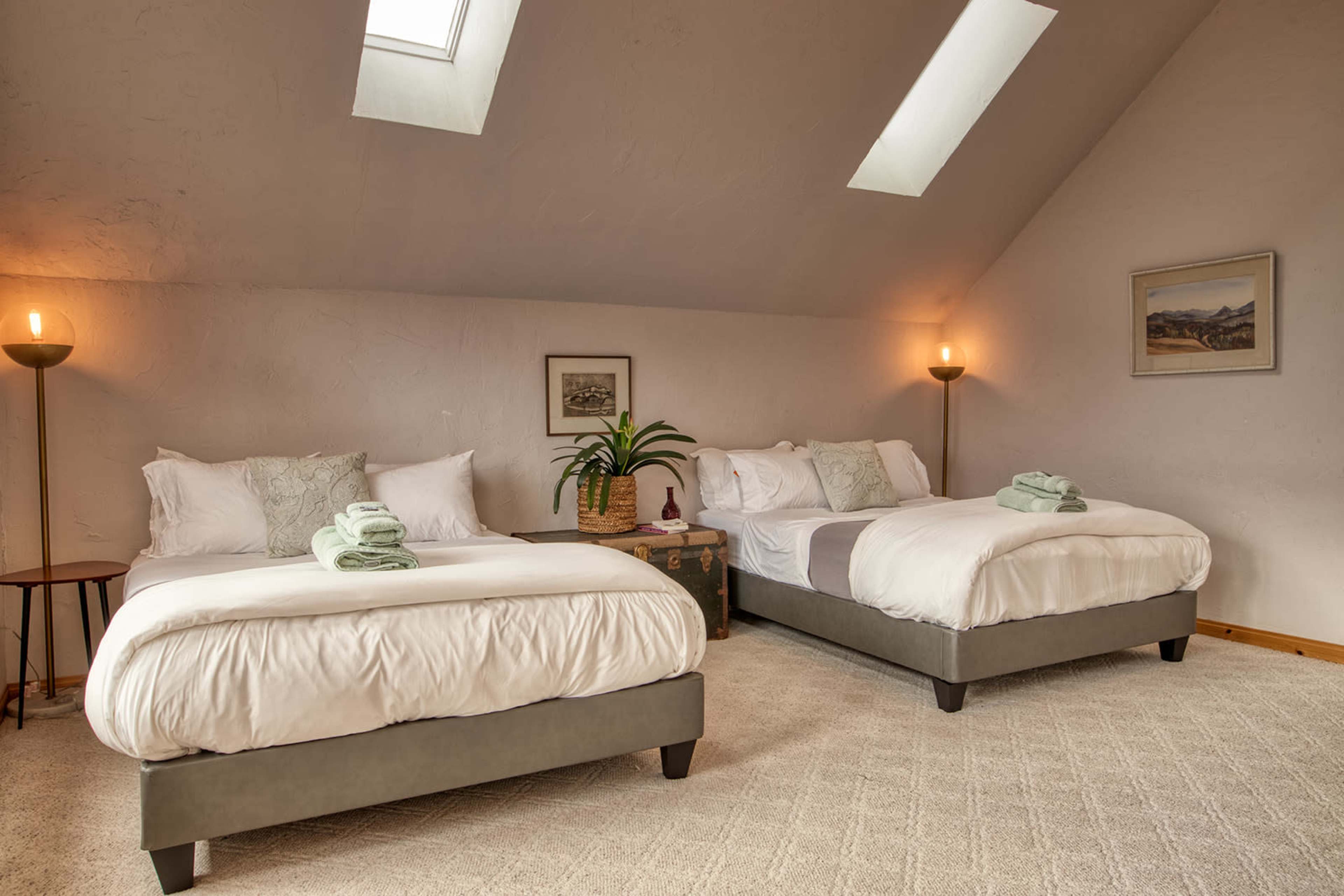 The image shows a bedroom with two large beds, each topped with green towels, a plant in the corner, and a skylight above.