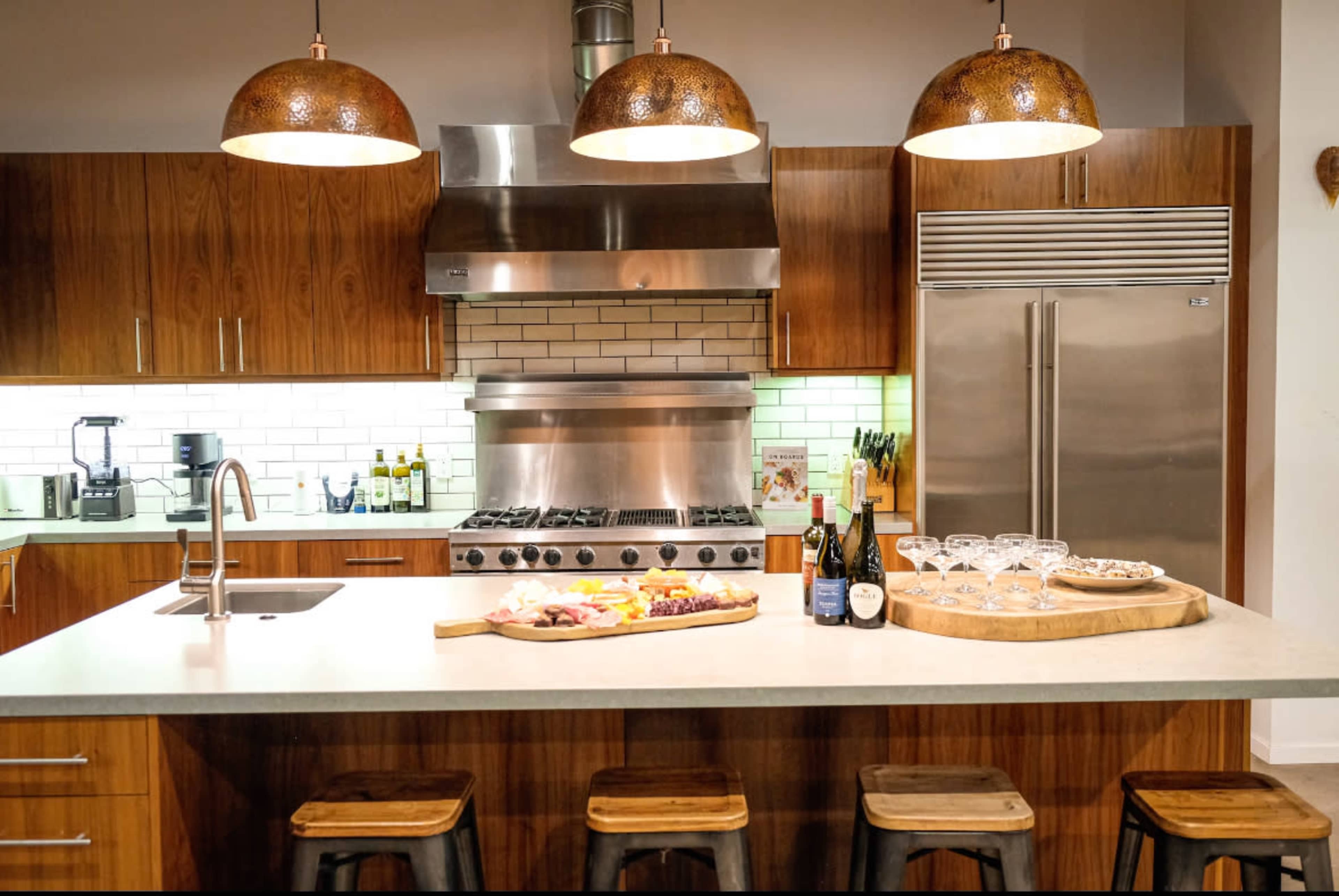 A modern kitchen features wooden cabinetry, stainless steel appliances, and a central island topped with a wooden platter of food and beverages, illuminated by decorative pendant lights.