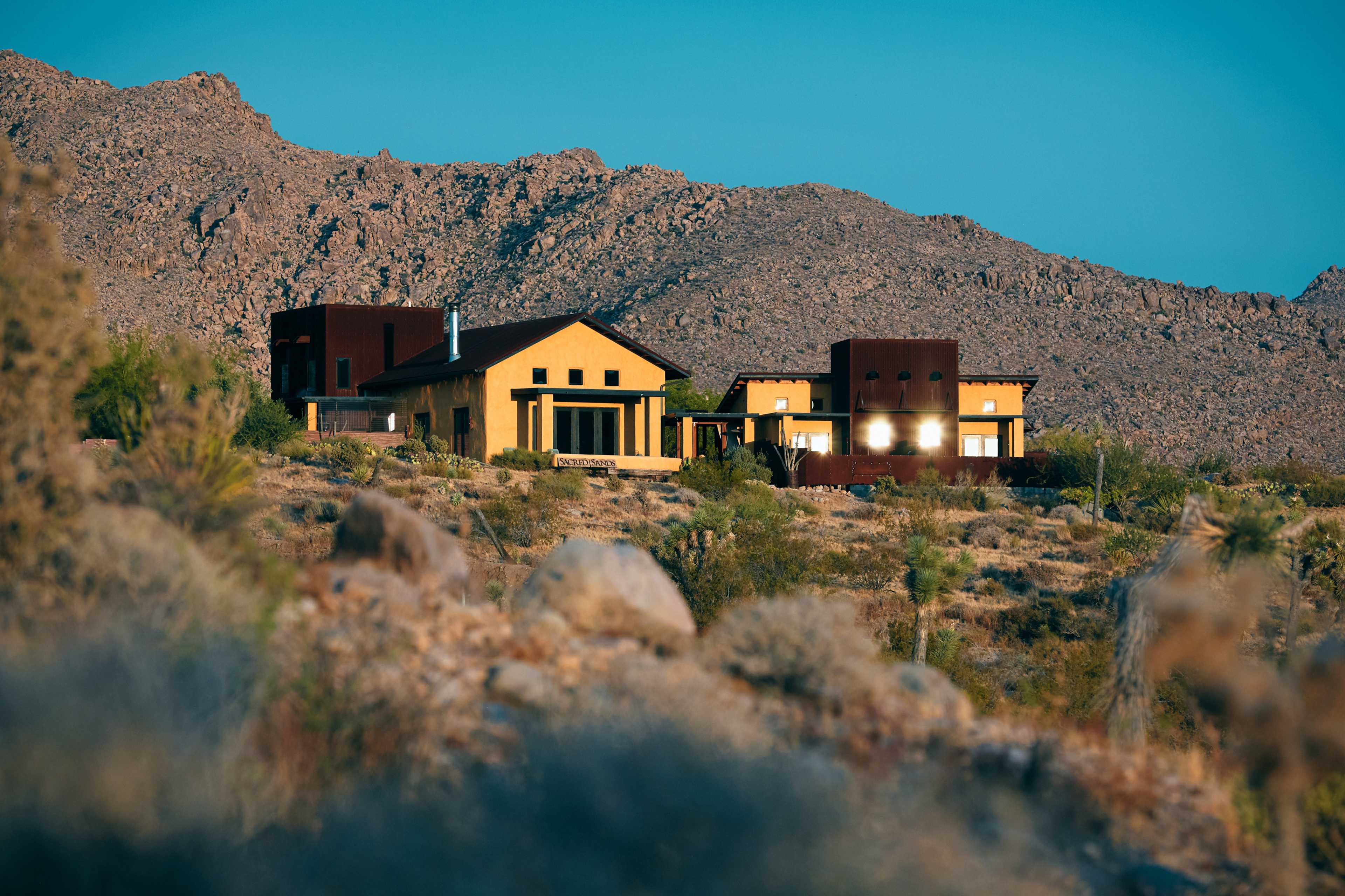 Two modern houses are nestled among rocky terrain and desert vegetation, set against a backdrop of rugged mountains.