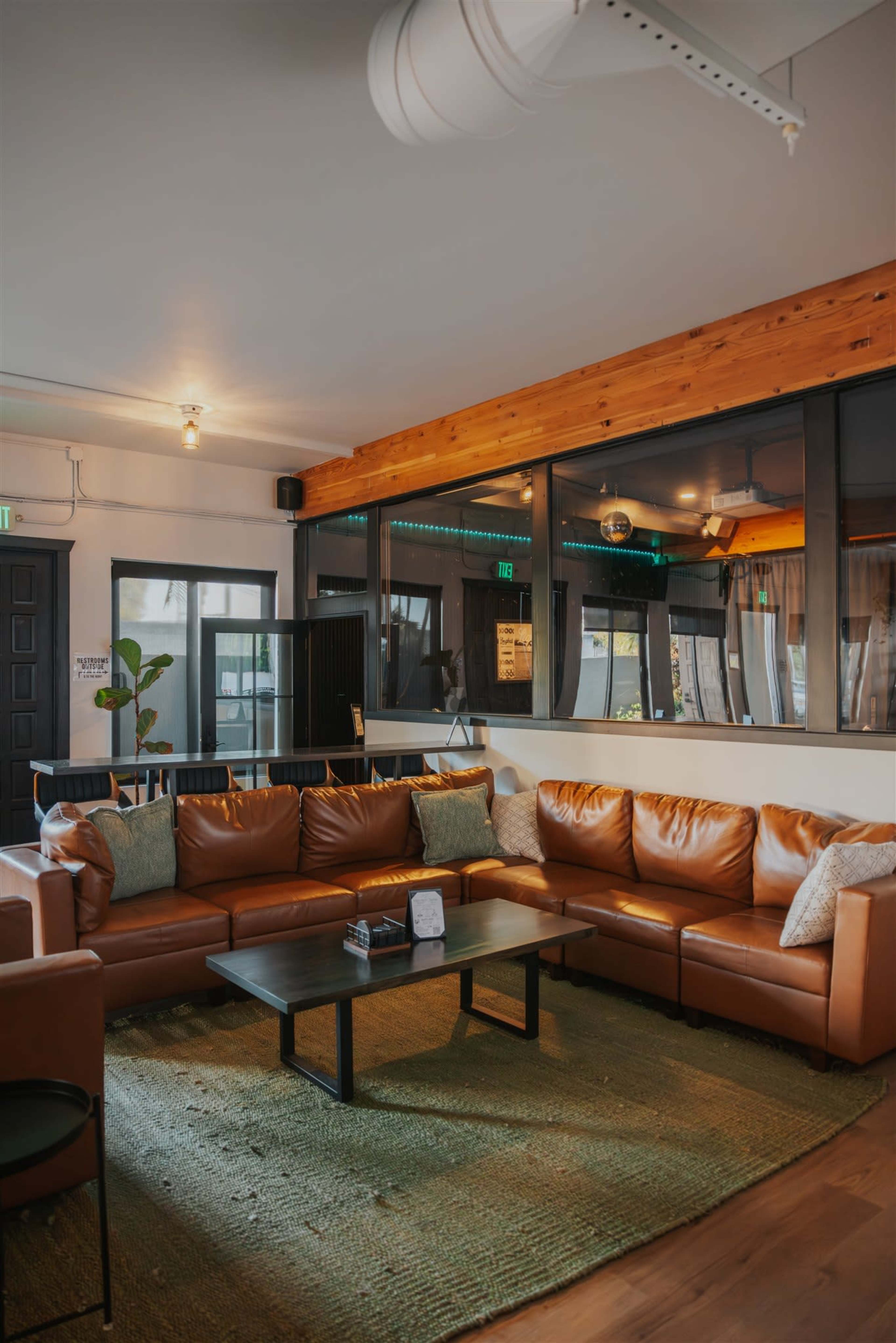 The image shows a cozy lounge area featuring a large brown leather sectional sofa, a black coffee table, and a green area rug, with large windows and wooden accents.