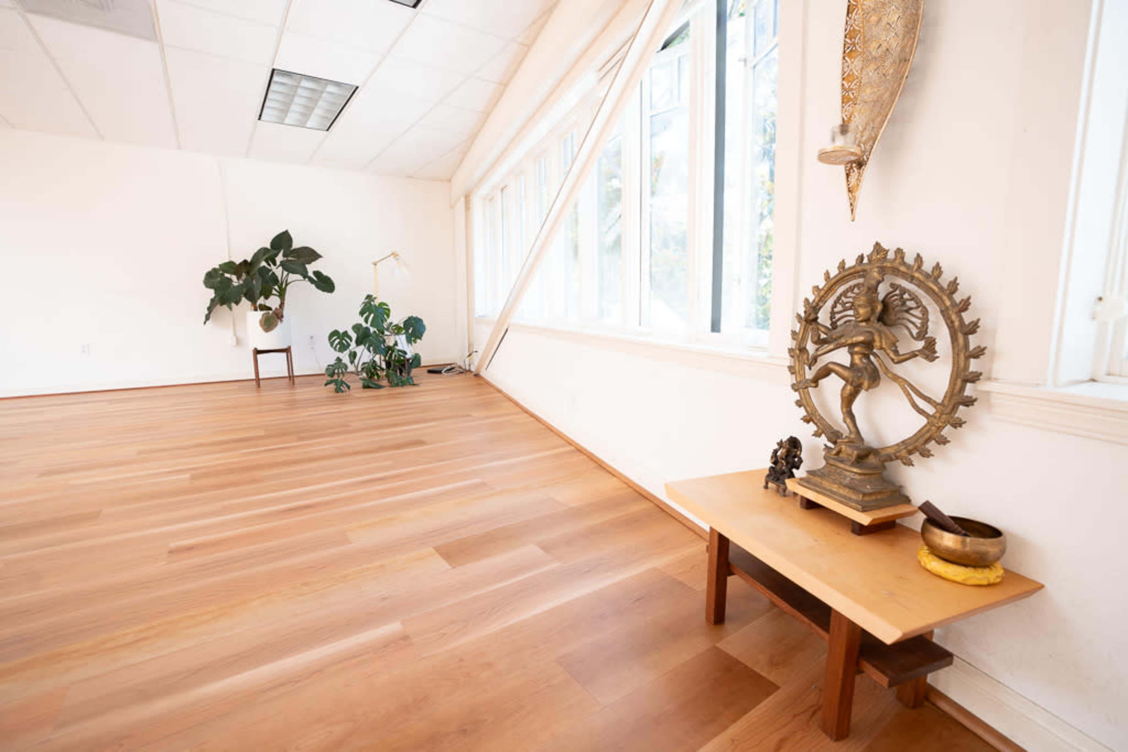 A spacious room with wooden flooring, large windows, and a decorative table featuring a brass statue and a bowl.