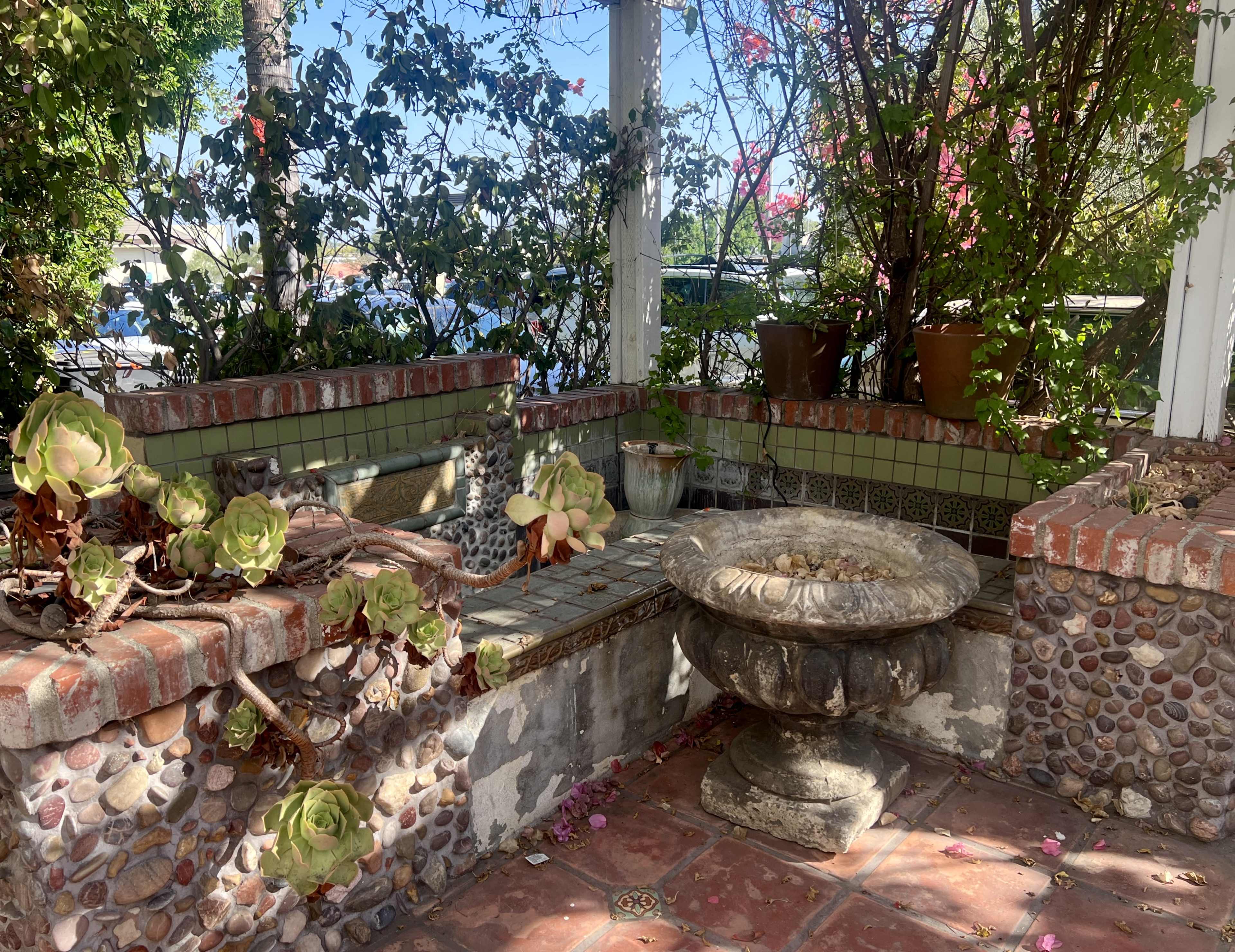 The image depicts a small outdoor garden area featuring a stone fountain surrounded by brick seating, potted plants, and succulents amidst a backdrop of lush greenery.
