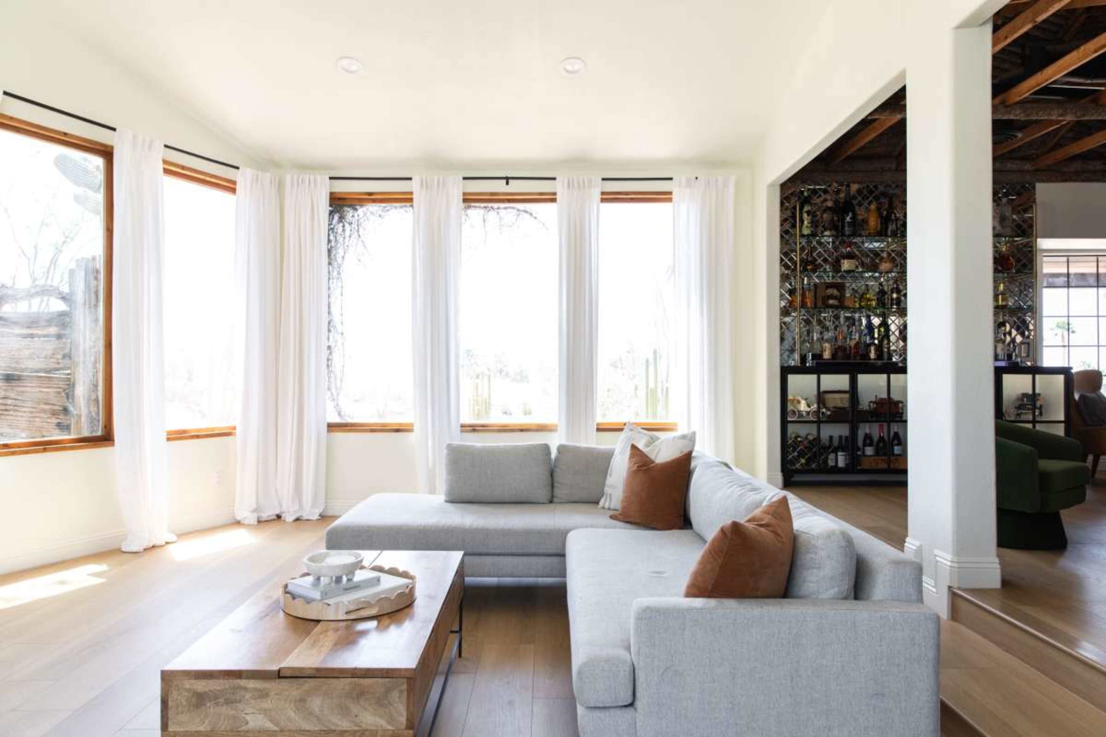 The image shows a modern living room with large windows, light curtains, a gray sectional sofa, and a wooden coffee table.