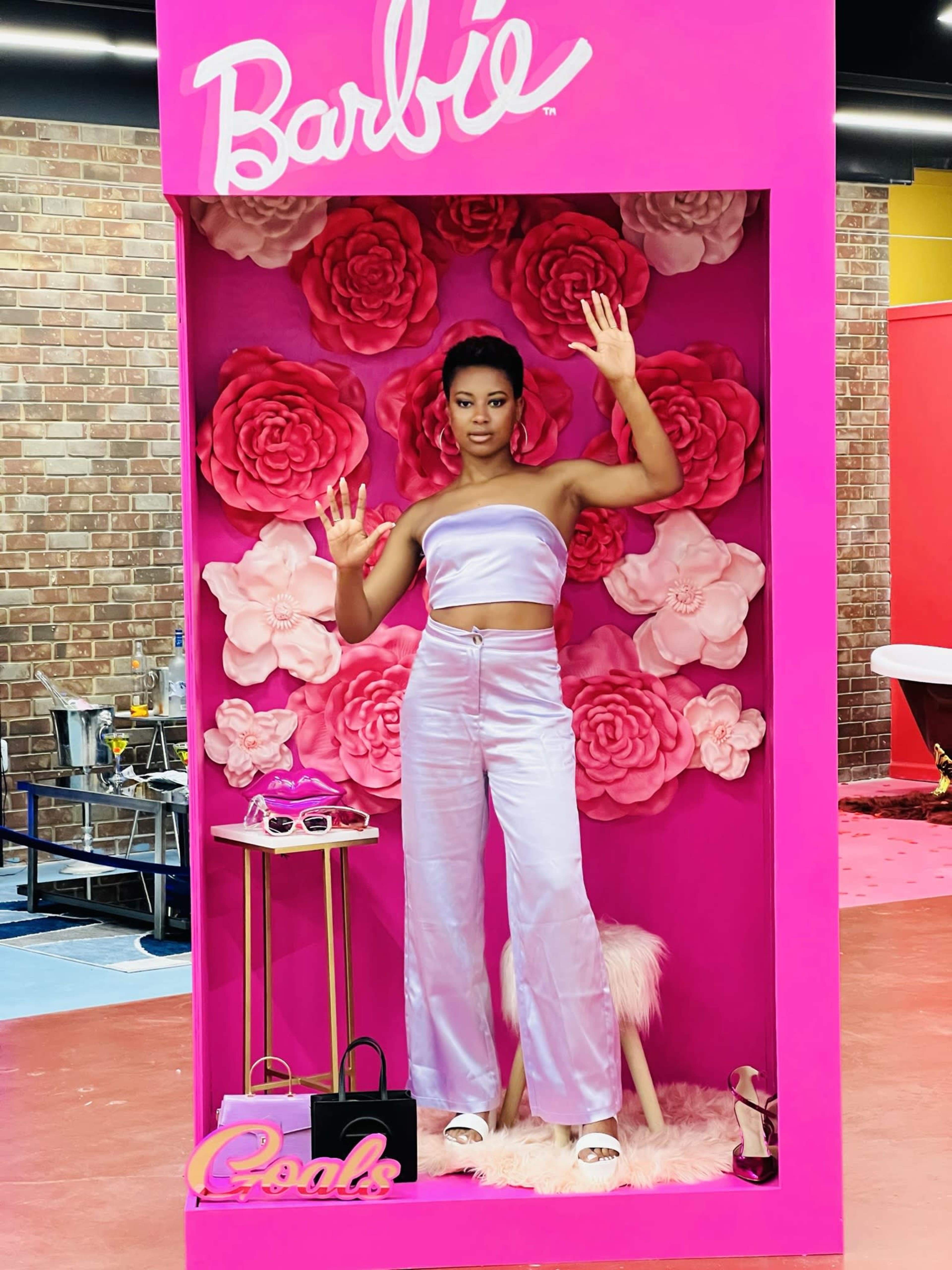 A model poses inside a pink display featuring large paper flowers and fashion accessories, wearing a lavender two-piece outfit.