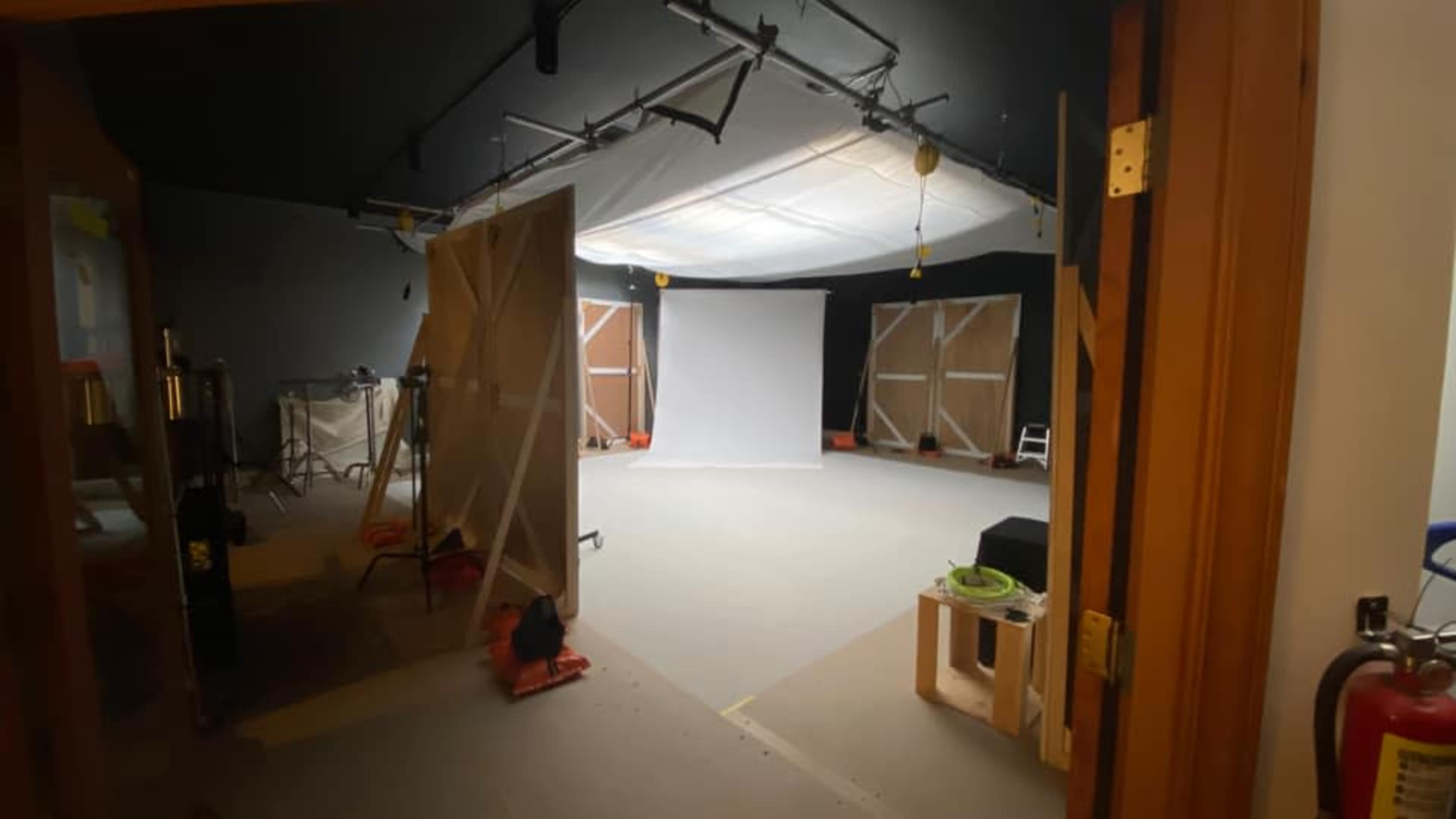 A photo studio with a large white backdrop in the center, surrounded by wooden panels and various equipment set up around the space.