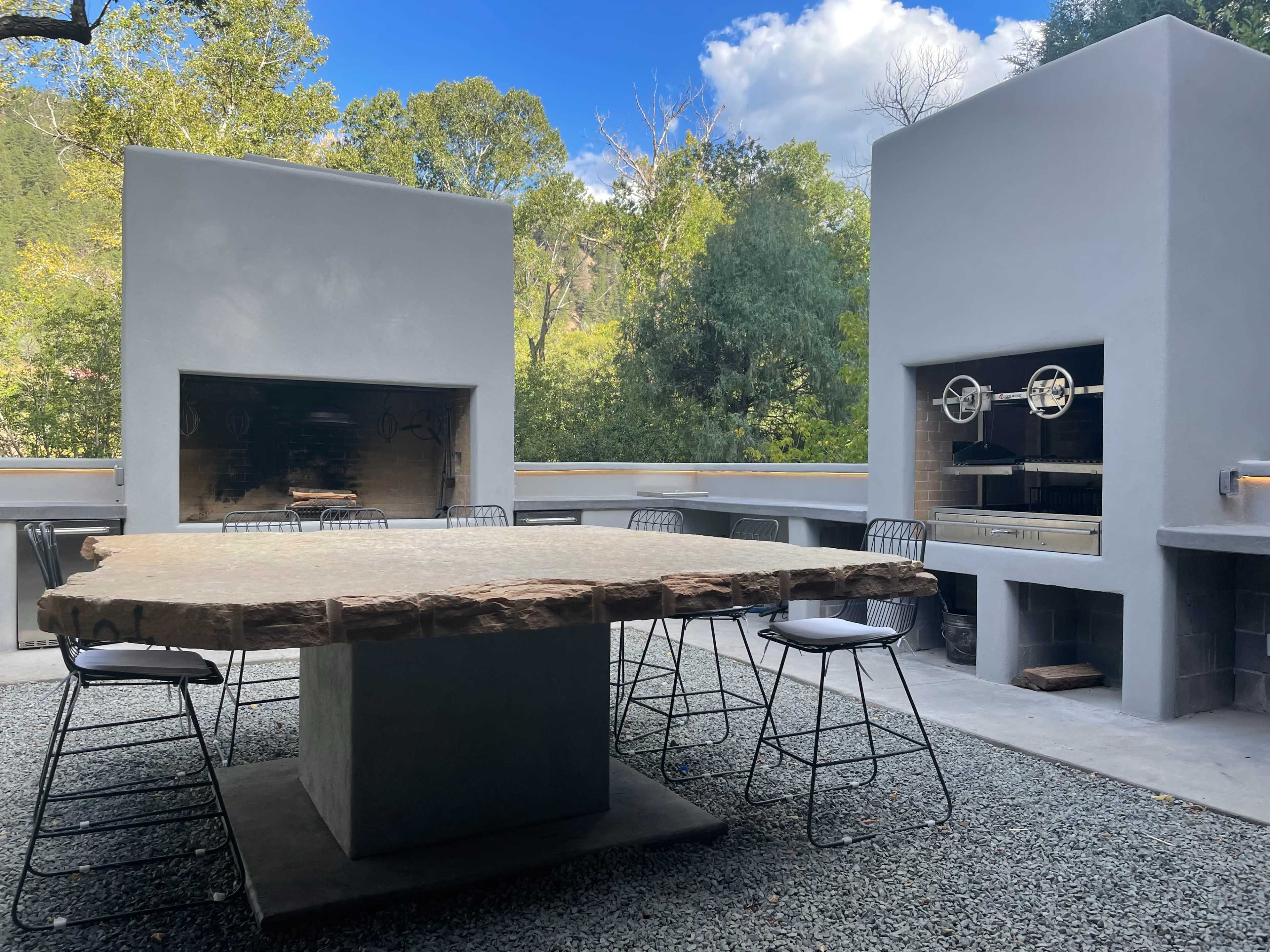 An outdoor kitchen area featuring a large stone dining table surrounded by metal chairs, with two built-in grilling stations and fireplaces positioned against white walls.