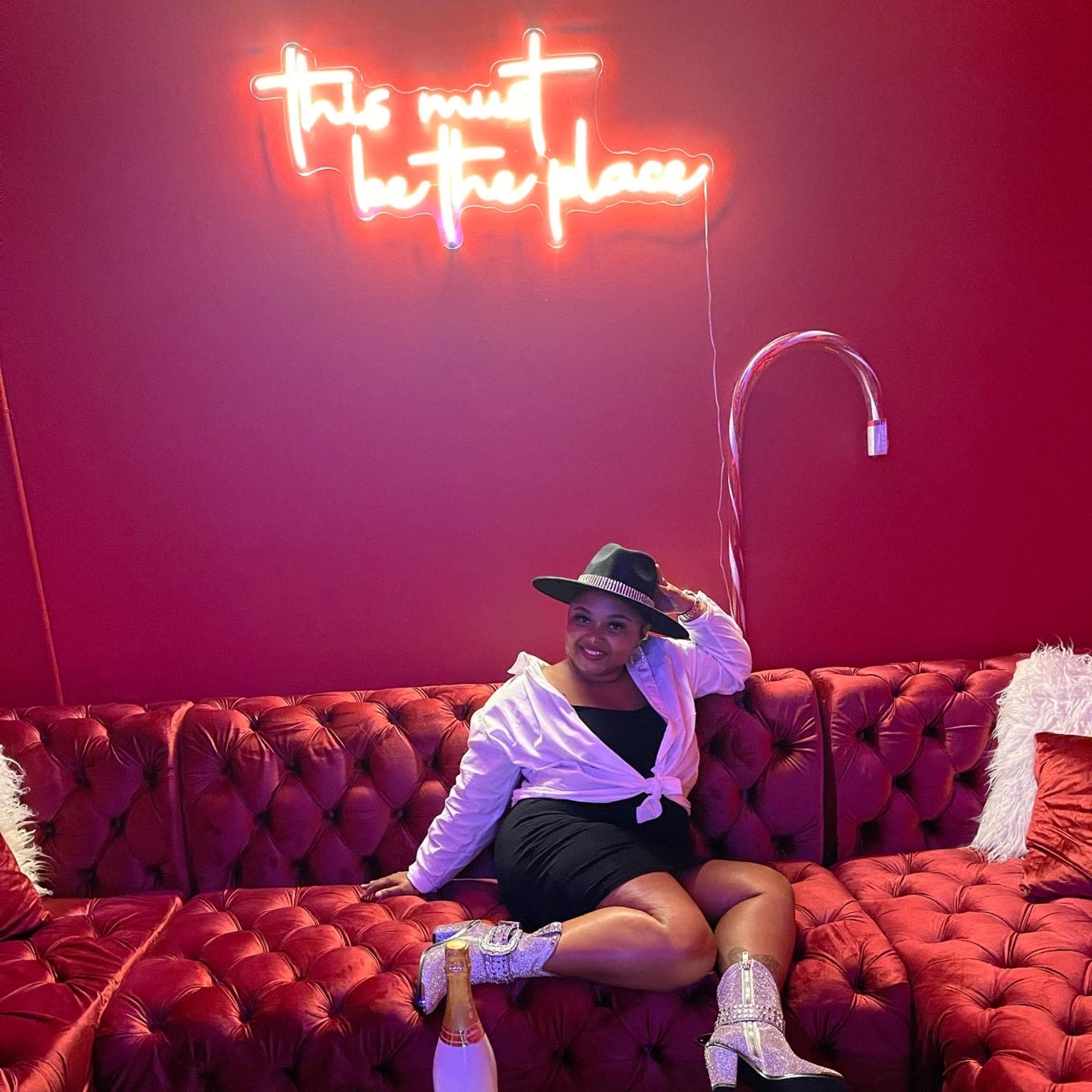 A person sits on a plush red couch under a neon sign that reads "this must be the place."