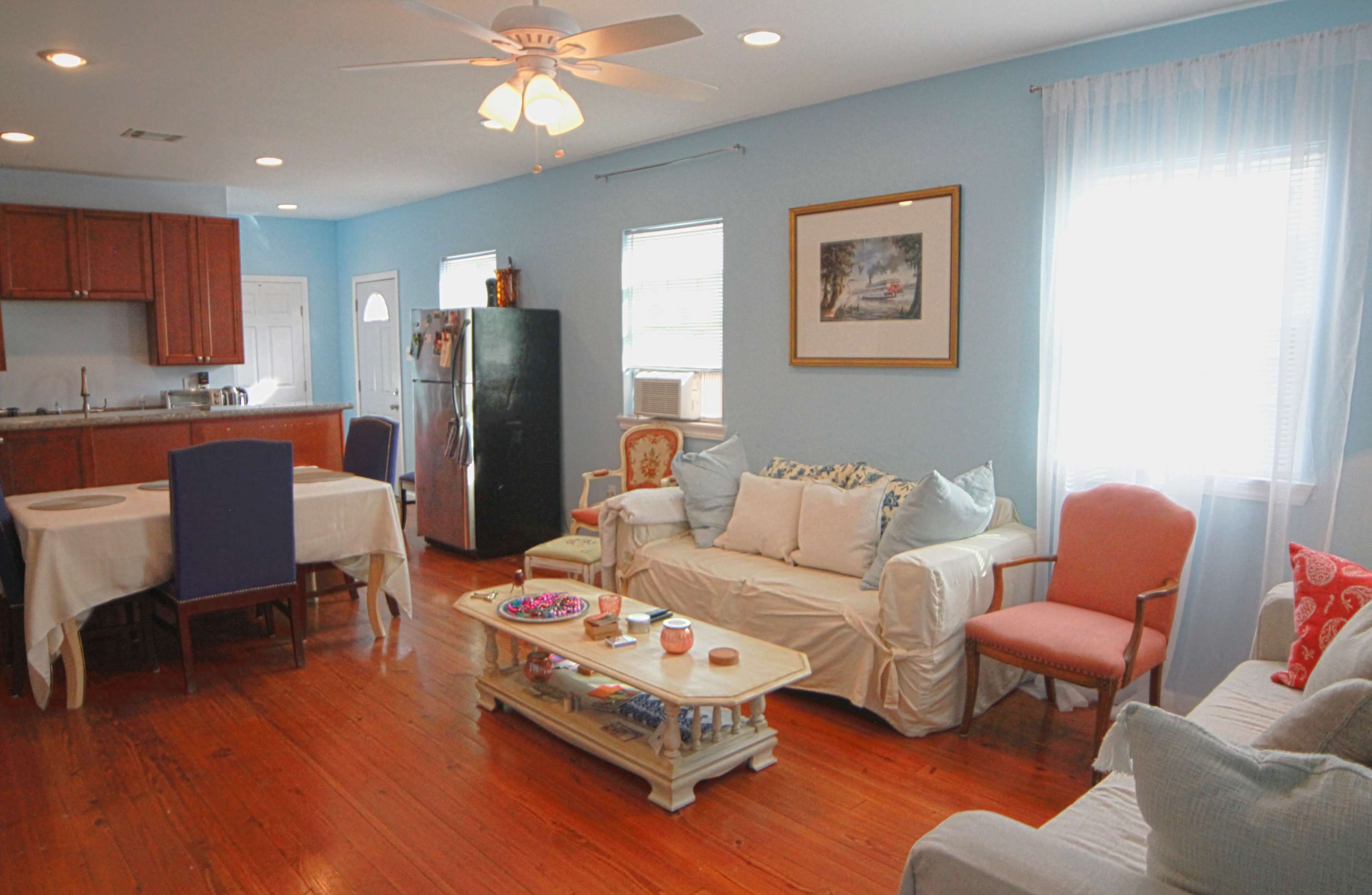 The image shows a brightly lit living area with a mix of wooden flooring, a white sofa, a dining table, and a kitchen visible in the background.